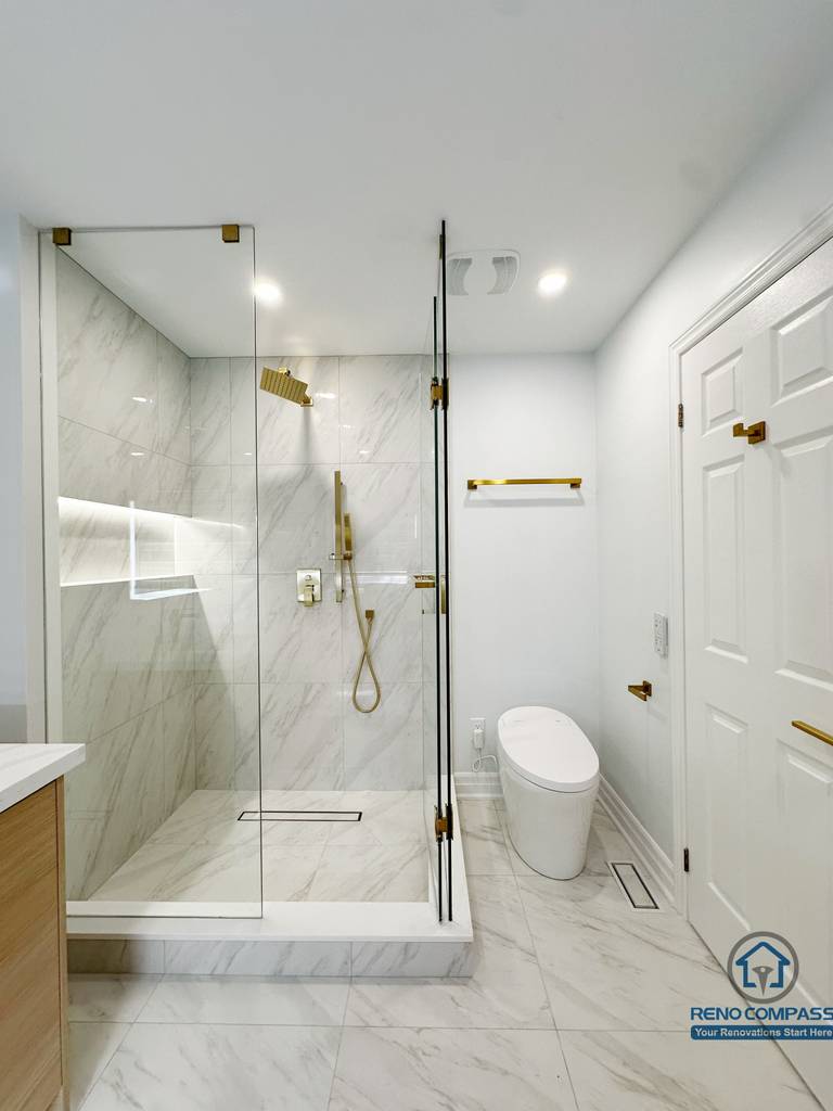 Contemporary bathroom renovation featuring a frameless glass walk-in shower, marble-look wall and floor tiles, built-in shower niche, and elegant gold-tone fixtures for a clean, modern finish.