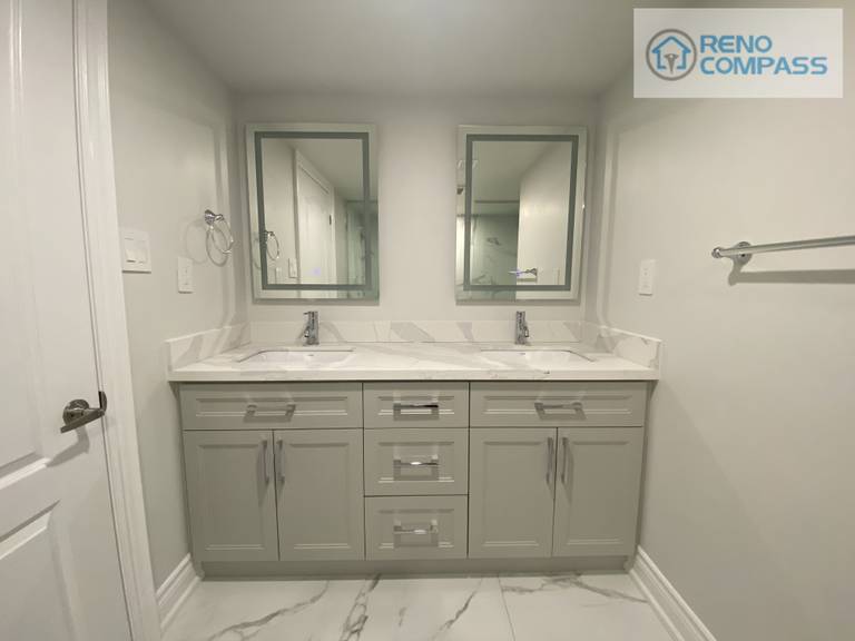 This Woodbridge basement bathroom renovation features a clean and functional double vanity design, ideal for shared use and daily convenience. The custom cabinetry provides ample storage while maintaining a streamlined appearance. A quartz countertop with subtle veining enhances durability and brightness, paired with integrated sinks and modern chrome fixtures for a cohesive look. Framed mirrors and neutral wall tones help reflect light and visually expand the space, making the basement bathroom feel open and comfortable. Completed by Reno Compass, a Toronto renovation contractor specializing in basement and bathroom renovations.