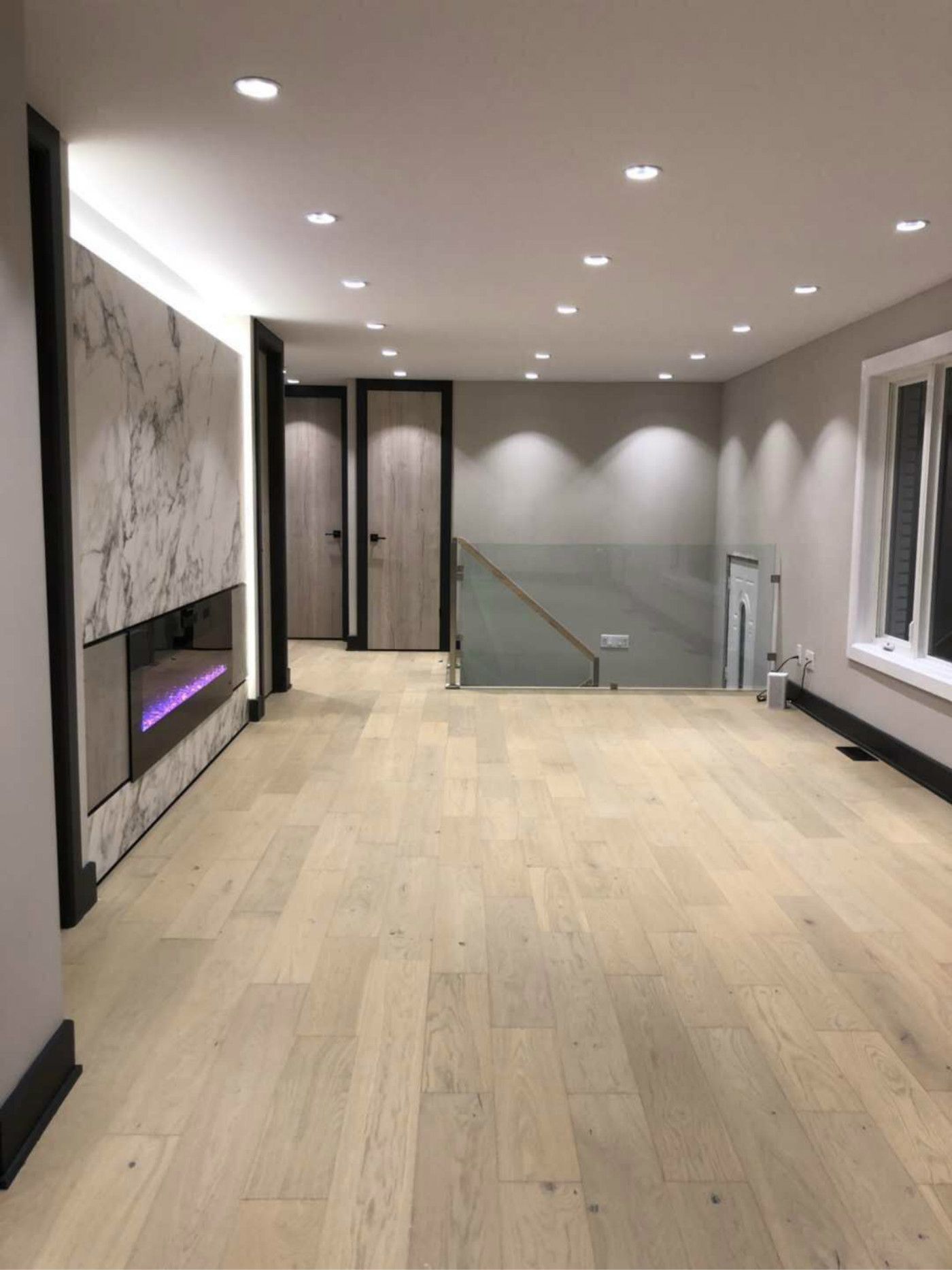 Professional basement finishing in Markham featuring wide-plank hardwood flooring, recessed pot lighting, marble-style feature wall, and a built-in linear electric fireplace with ambient lighting. The open layout with glass railing staircase and modern interior doors creates a bright, spacious entertainment area ideal for family gatherings and added home value.