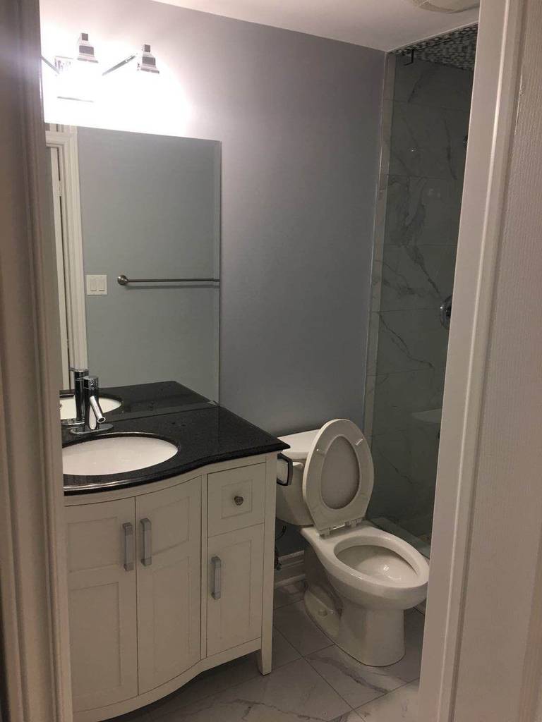 Completed basement bathroom as part of a Toronto basement renovation project. This space includes a modern white vanity cabinet with black countertop, under-mount sink, updated lighting fixture, full-height mirror, standard toilet installation, and a glass-enclosed shower with marble-style wall tiles. The layout maximizes functionality while maintaining a clean, contemporary basement bathroom design.