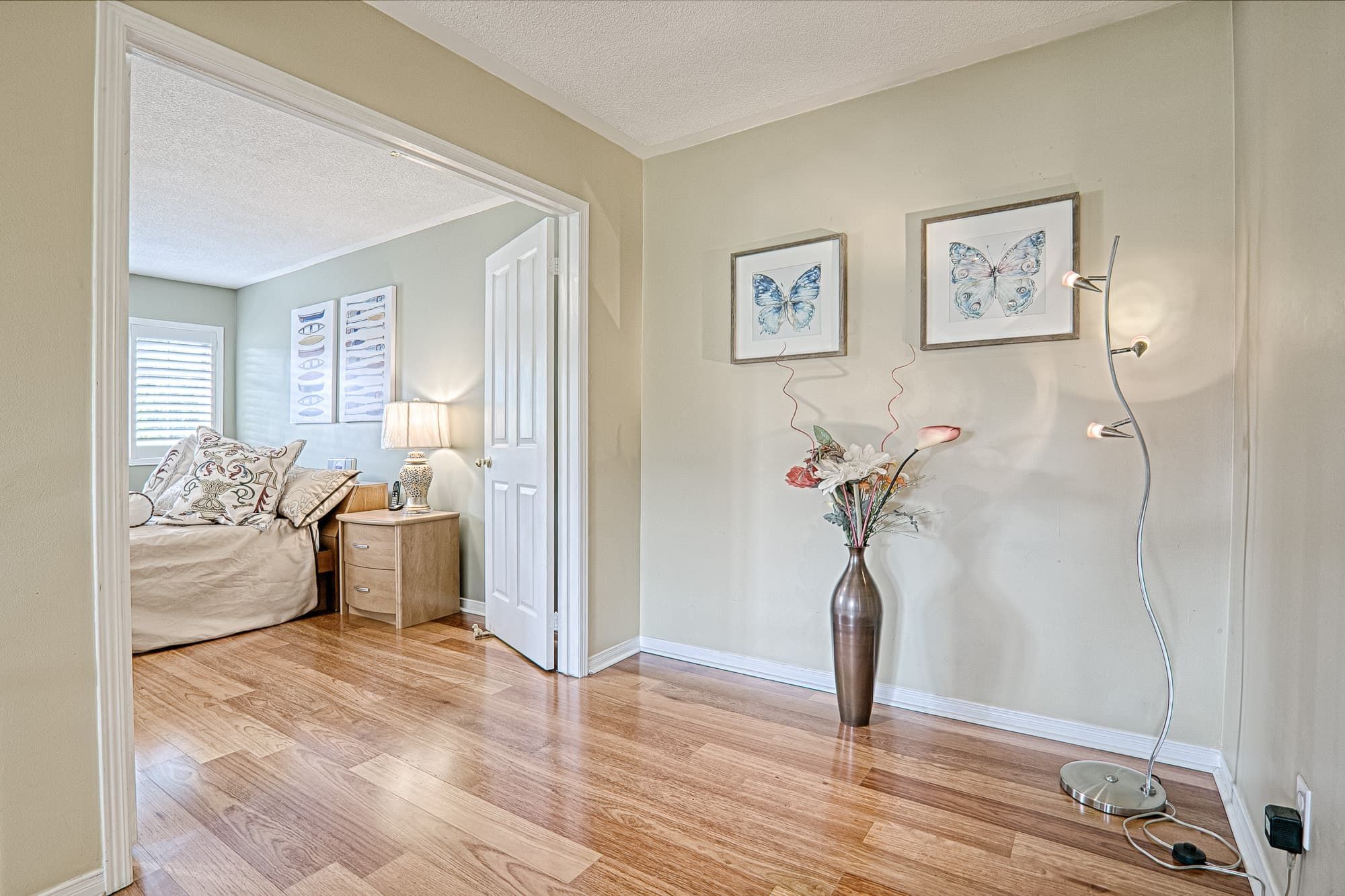 This open bedroom entrance and hallway space highlights warm hardwood flooring, soft beige wall tones, and simple decorative accents that create a seamless and inviting transition between rooms. The natural light, coordinated bedside furniture, and modern floor lamp enhance both comfort and functionality. A refined example of warm minimalist interior renovation and cohesive home styling for homeowners in Toronto and the GTA.
