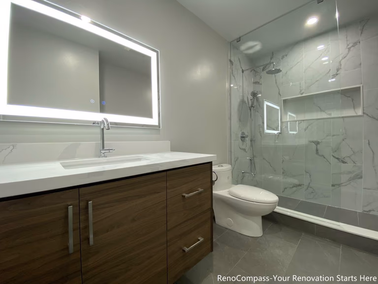 This Oakville bathroom renovation showcases a modern walk-in glass shower with marble-look wall tiles, built-in shower niche, and sleek frameless enclosure for a clean contemporary design. The space also features a custom wood vanity with quartz countertop, LED backlit mirror, and upgraded fixtures that enhance both functionality and visual appeal. Completed by a professional Toronto renovation company, this bathroom upgrade focuses on maximizing brightness, storage, and comfort while delivering a stylish and durable modern bathroom space.