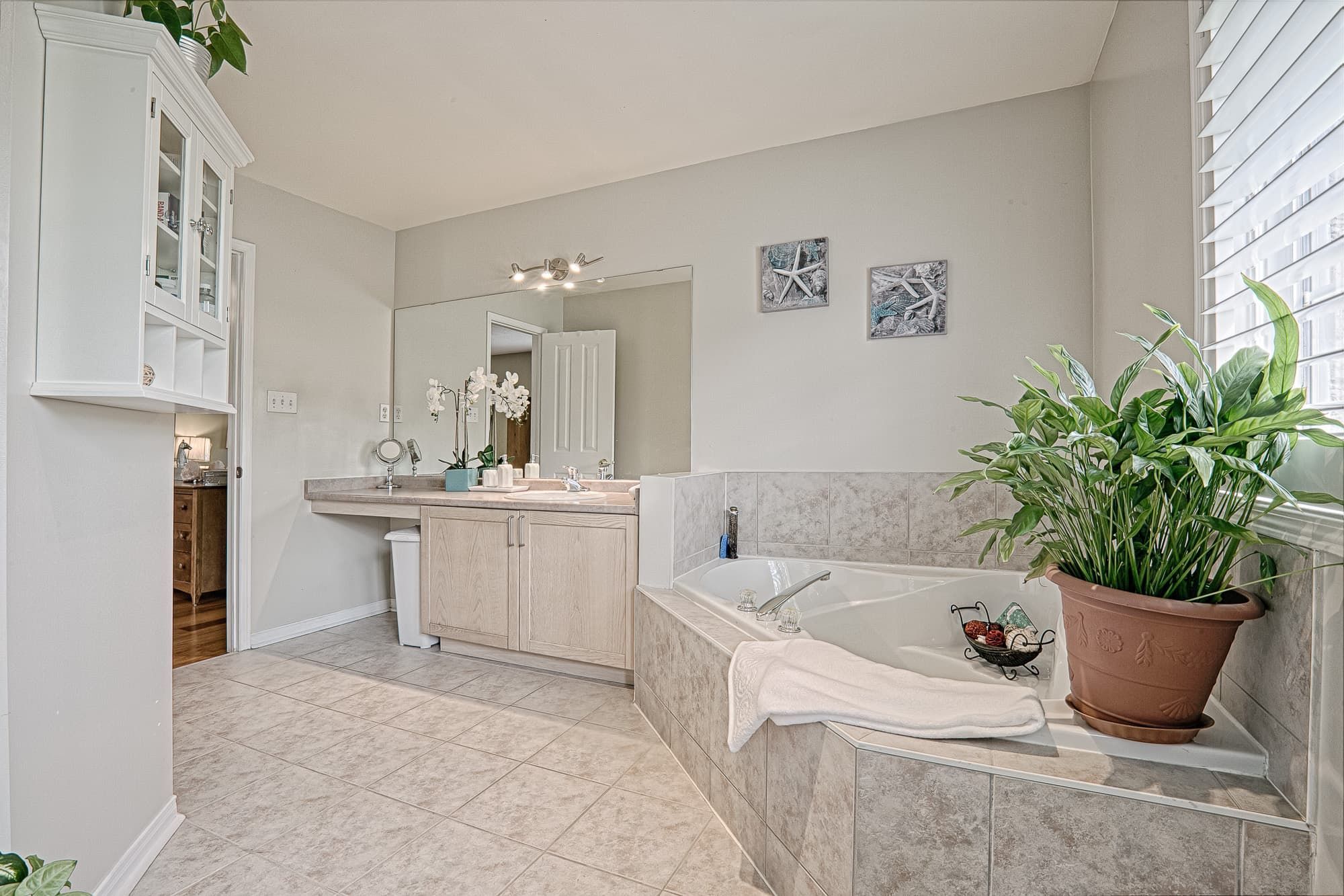 This warm minimalist bathroom design features a spacious built-in soaking tub, large vanity with extended countertop, and soft neutral tile finishes that create a relaxing and elegant atmosphere. The oversized mirror enhances natural light, while subtle décor elements and indoor plants add warmth and balance to the space. A perfect example of modern, cozy bathroom interior design for homeowners seeking functional yet stylish renovation ideas in Toronto and the GTA.