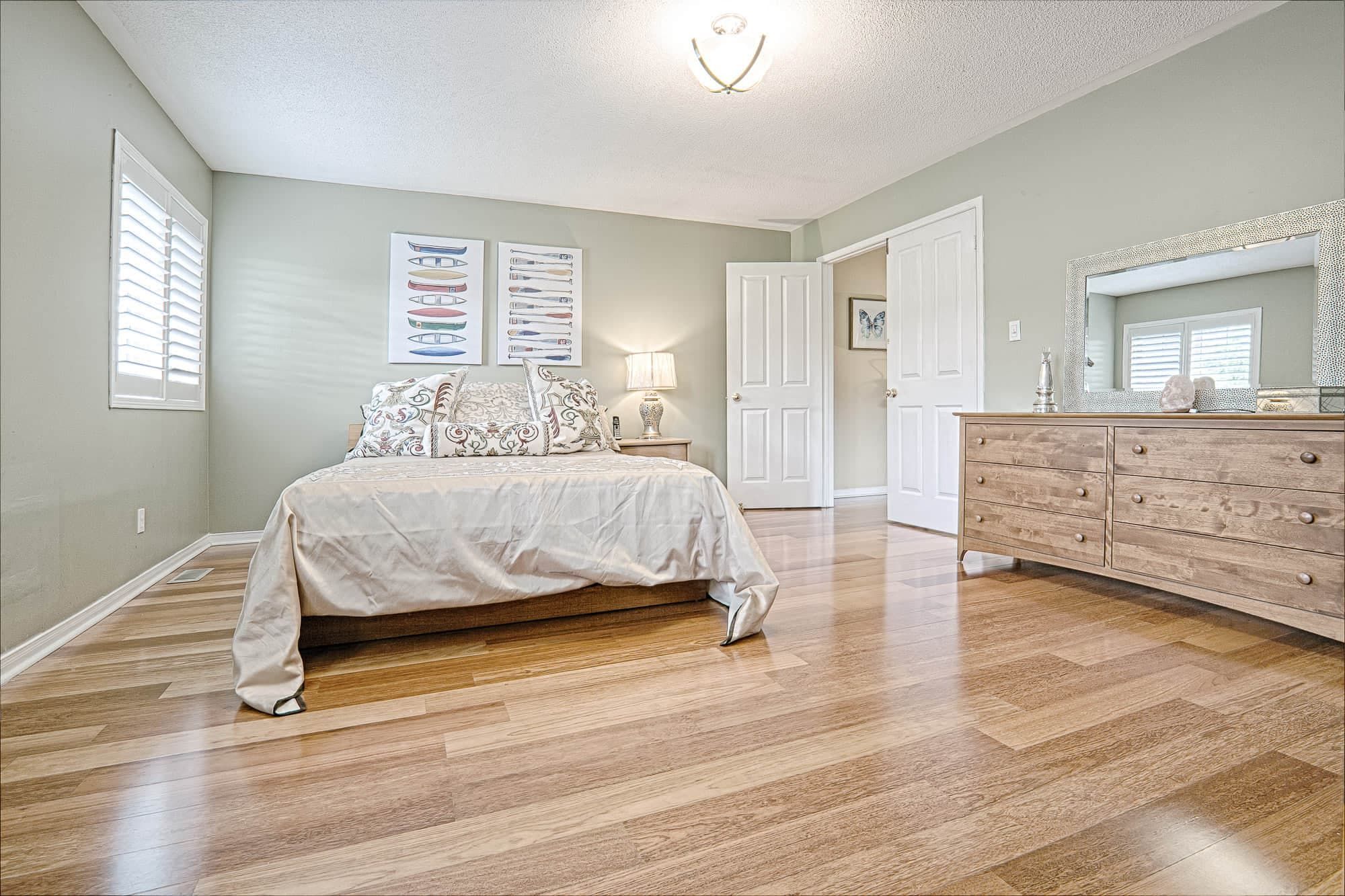 This warm minimalist bedroom interior features soft neutral wall tones, natural hardwood flooring, and clean-lined furniture that create a bright and inviting atmosphere. The spacious layout, modern dresser with framed mirror, and simple yet elegant bedding design reflect a balanced combination of comfort and functionality. Ideal inspiration for homeowners looking for cozy interior design and modern home styling ideas in Toronto and the GTA.