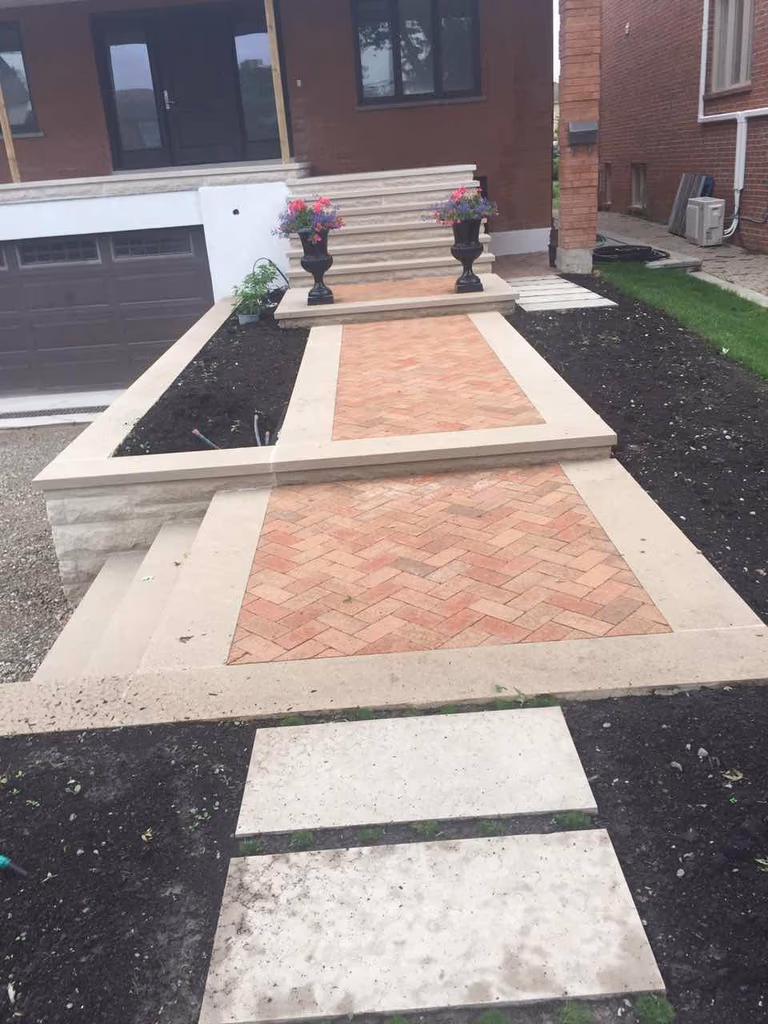 This Toronto interlocking walkway showcases a stylish herringbone brick pattern framed with natural stone borders, leading to a raised front entrance with elegant steps. Combined with fresh garden beds and decorative planters, the design enhances curb appeal while creating a welcoming and functional outdoor space.