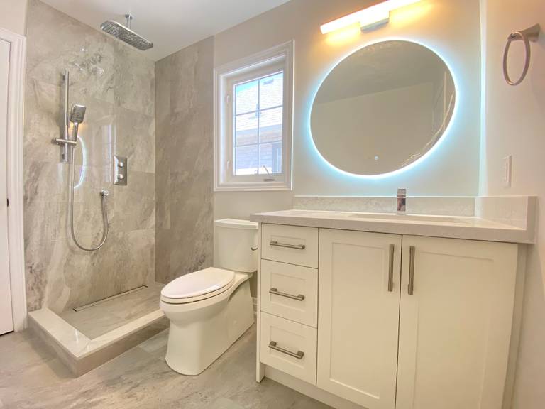 A modern secondary bathroom completed as part of a Richmond Hill interior renovation. This bathroom renovation features a walk-in shower with large-format tile walls, a compact vanity with integrated storage, an LED backlit round mirror, and neutral tile flooring for a clean and bright finish.