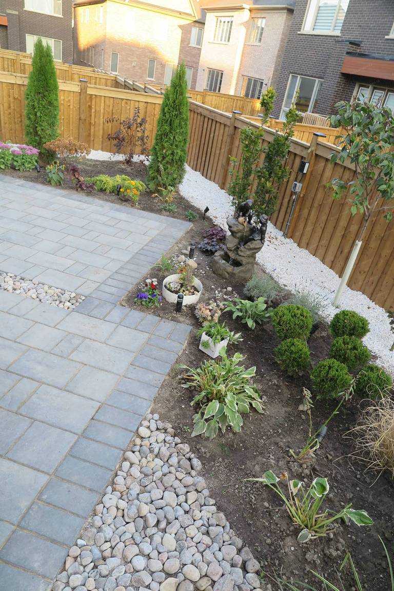 Backyard landscaping renovation in Vaughan featuring a modern interlocking patio, decorative river rock accents, layered garden beds, and a small ornamental water feature. This outdoor design blends hardscape and soft scape elements to create a relaxing backyard environment with strong visual balance and natural landscape texture.