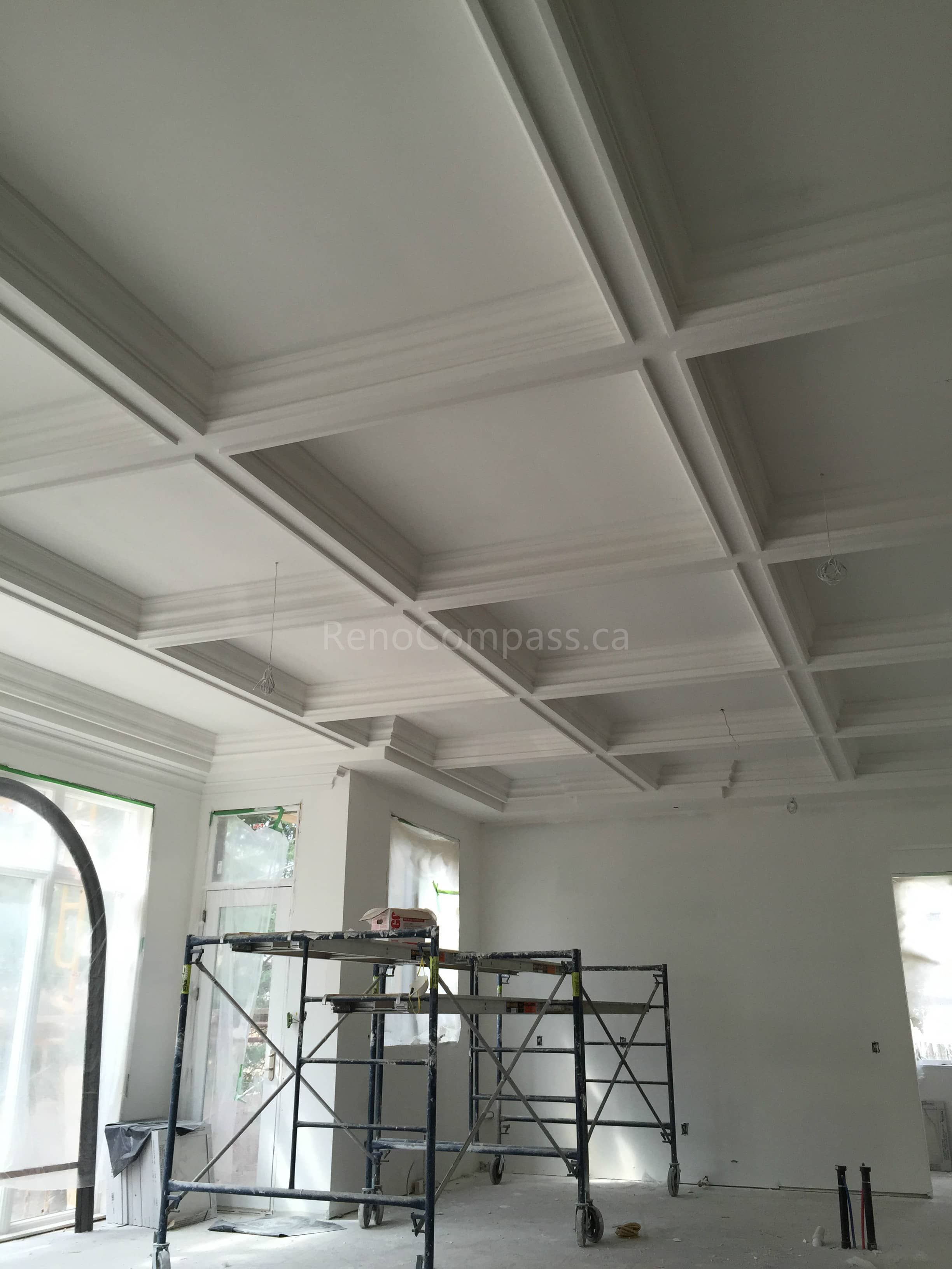 This Toronto interior renovation project features a custom coffered ceiling installation in progress, highlighting detailed trim work and precision framing. The elegant ceiling grid design adds depth, character, and architectural interest to the living space. Large windows allow natural light to enhance the craftsmanship and overall openness of the room. Completed by Reno Compass Inc., this project showcases professional ceiling finishing and high-end interior renovation services in Toronto.