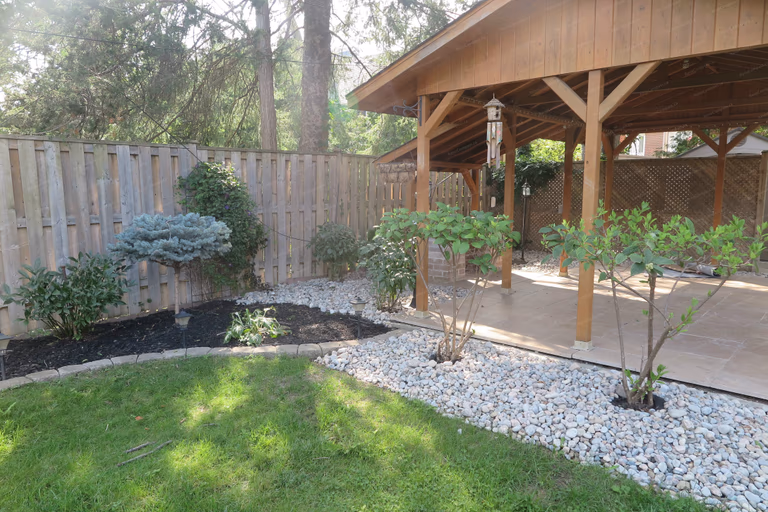 North York outdoor landscaping project showcasing decorative white gravel garden beds, newly planted shrubs, natural lawn area, and a custom wooden pavilion. Designed and built by Reno Compass to create a clean, low-maintenance backyard with a relaxing outdoor living and garden space.