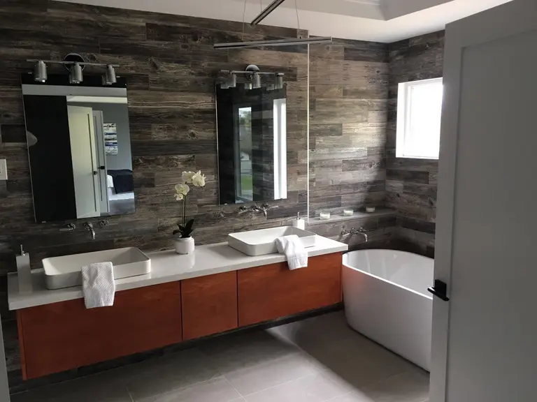 This Toronto interior renovation project showcases a modern bathroom transformation featuring a floating wood vanity with double vessel sinks, sleek wall-mounted mirrors, and a freestanding soaking tub. The textured wood accent wall adds warmth and depth, while large-format flooring enhances the clean, contemporary aesthetic. Designed for both functionality and luxury, this bathroom renovation reflects high-quality craftsmanship and modern interior styling by a professional Toronto renovation company.