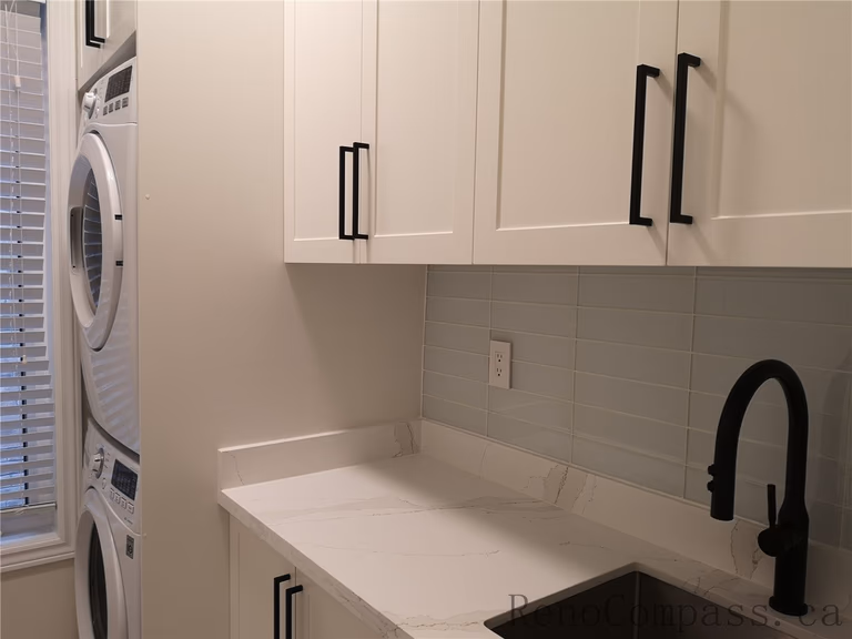 This Aurora interior renovation features a modern laundry room with custom white shaker cabinets, quartz countertop, and integrated utility sink with matte black faucet. The stacked washer and dryer design maximizes space while maintaining a clean and functional layout. Completed by a professional Toronto renovation company focused on efficient storage solutions, modern finishes, and high-quality craftsmanship for everyday living.