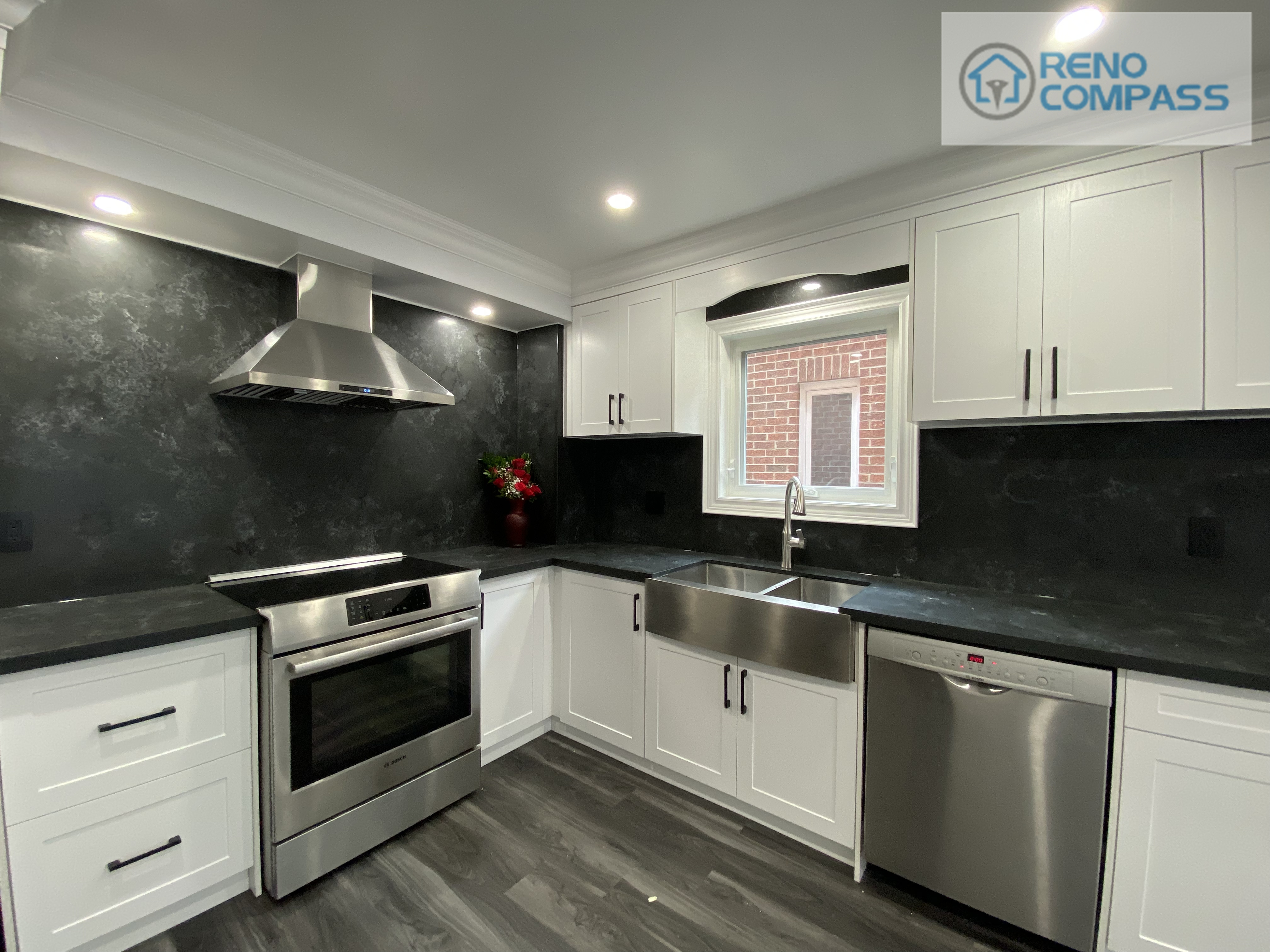 Wide-angle view of a modern kitchen interior renovation in Pickering, featuring full-height white shaker cabinetry, black stone countertops and backsplash, stainless steel appliances, recessed ceiling lighting, and durable vinyl plank flooring. This clean and functional kitchen layout was designed and built by Reno Compass Inc., delivering a bright, streamlined space optimized for everyday use.
