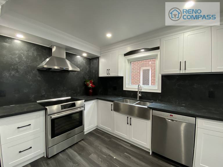 Wide-angle view of a modern kitchen interior renovation in Pickering, featuring full-height white shaker cabinetry, black stone countertops and backsplash, stainless steel appliances, recessed ceiling lighting, and durable vinyl plank flooring. This clean and functional kitchen layout was designed and built by Reno Compass Inc., delivering a bright, streamlined space optimized for everyday use.