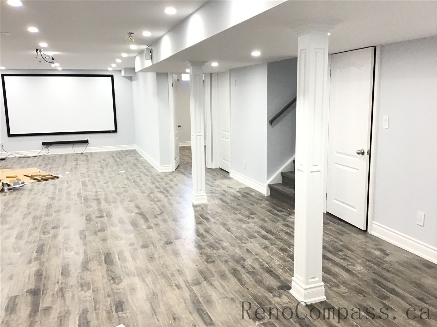 Spacious open-concept basement renovation in Richmond Hill featuring durable vinyl plank flooring, recessed pot lights, structural support columns, and a large wall-mounted projector screen for a home theater setup. Designed for entertainment and functionality, this finished basement showcases clean modern finishes completed by a trusted Toronto Renovation Company.