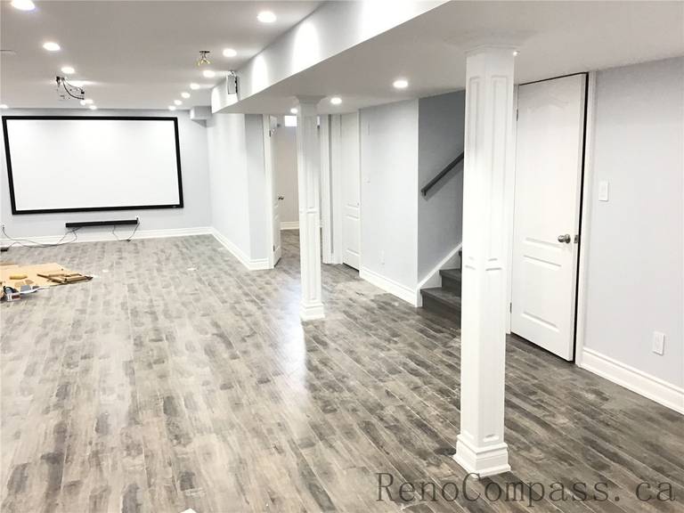 Spacious open-concept basement renovation in Richmond Hill featuring durable vinyl plank flooring, recessed pot lights, structural support columns, and a large wall-mounted projector screen for a home theater setup. Designed for entertainment and functionality, this finished basement showcases clean modern finishes completed by a trusted Toronto Renovation Company.