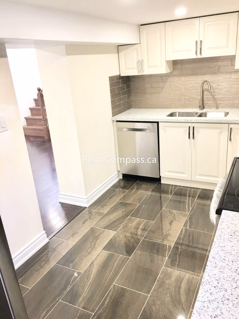 This Toronto basement renovation showcases a modern kitchenette with custom white shaker cabinets, quartz countertop, and a stylish textured backsplash. The compact layout includes a double sink, built-in microwave space, and durable tile flooring, creating a clean and functional secondary kitchen. Completed by a trusted Toronto renovation company, this basement kitchen upgrade adds convenience, value, and contemporary appeal to the home.