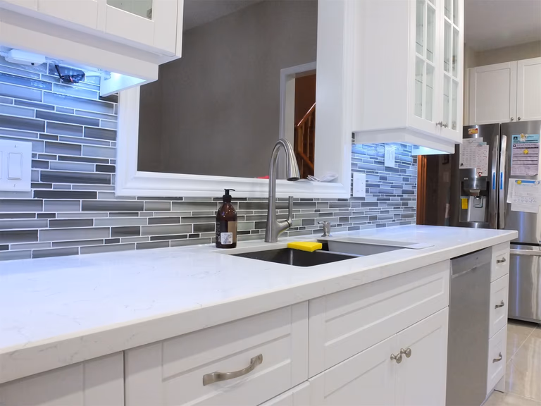This renovated kitchen in Vaughan features a modern undermount sink, sleek quartz countertops, and white shaker cabinets. The stylish glass mosaic backsplash combined with LED under-cabinet lighting adds depth and elegance to the space. Completed by a professional Toronto renovation company, the design enhances both practicality and contemporary kitchen aesthetics.
