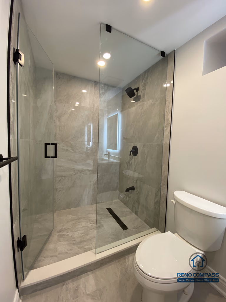 This Markham bathroom renovation features a modern walk-in shower with frameless glass panels and large-format marble-style porcelain tiles. The extended shower layout improves daily usability while maintaining a clean, open visual flow. Black shower fixtures, a linear floor drain, and full-height wall tiling create a refined, contemporary look with strong durability and easy maintenance. Designed and built by a professional Toronto renovation company, this bathroom upgrade balances functionality, waterproof performance, and long-term aesthetic value for residential homes.
