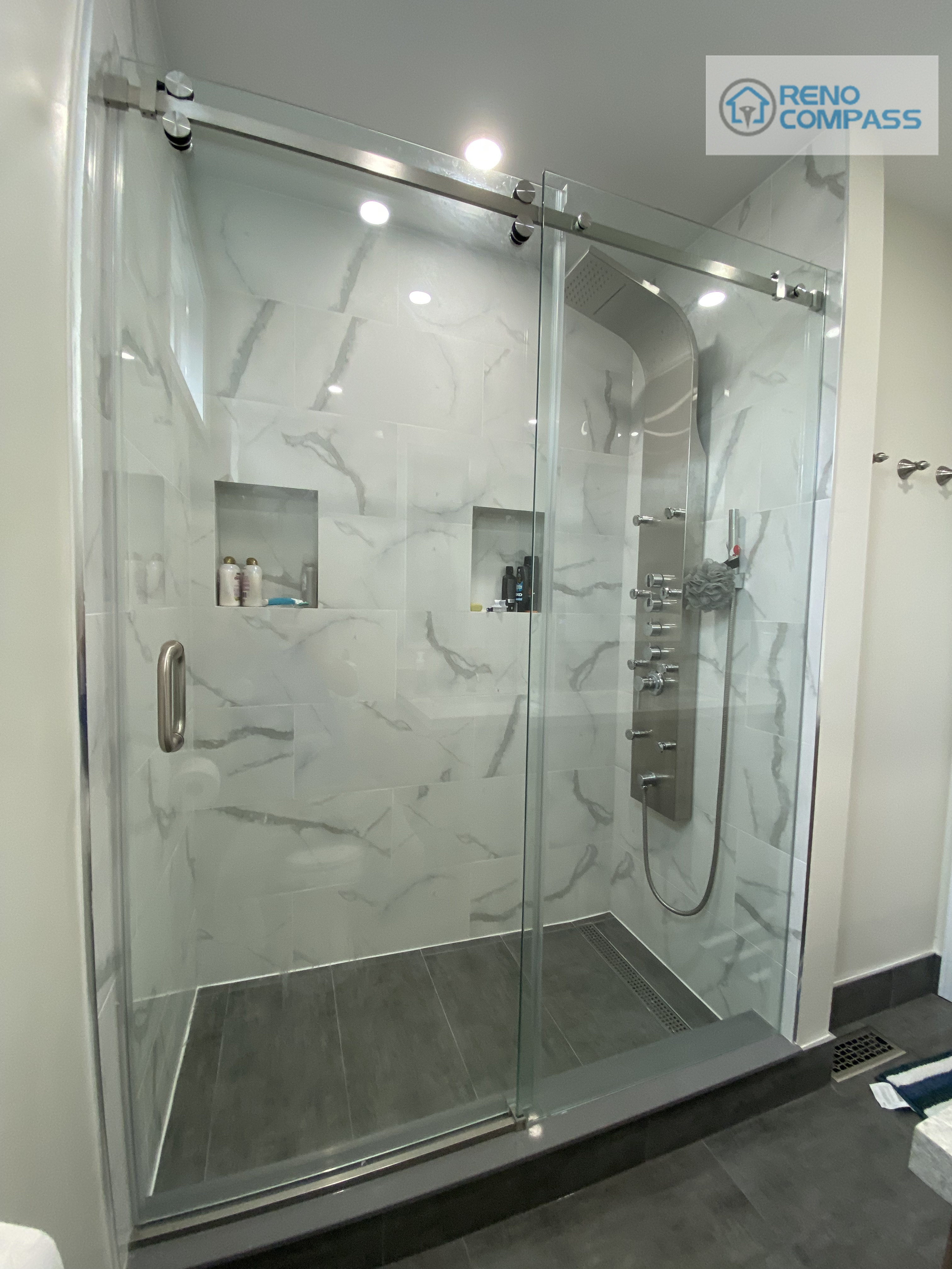 This bathroom renovation in Pickering features a frameless glass walk-in shower with marble-look wall tiles, built-in shower niches, sliding glass door system, and modern rainfall shower fixtures. The clean, hotel-style design emphasizes functionality, waterproof detailing, and contemporary aesthetics, completed by Reno Compass Inc. as part of a full interior renovation project.