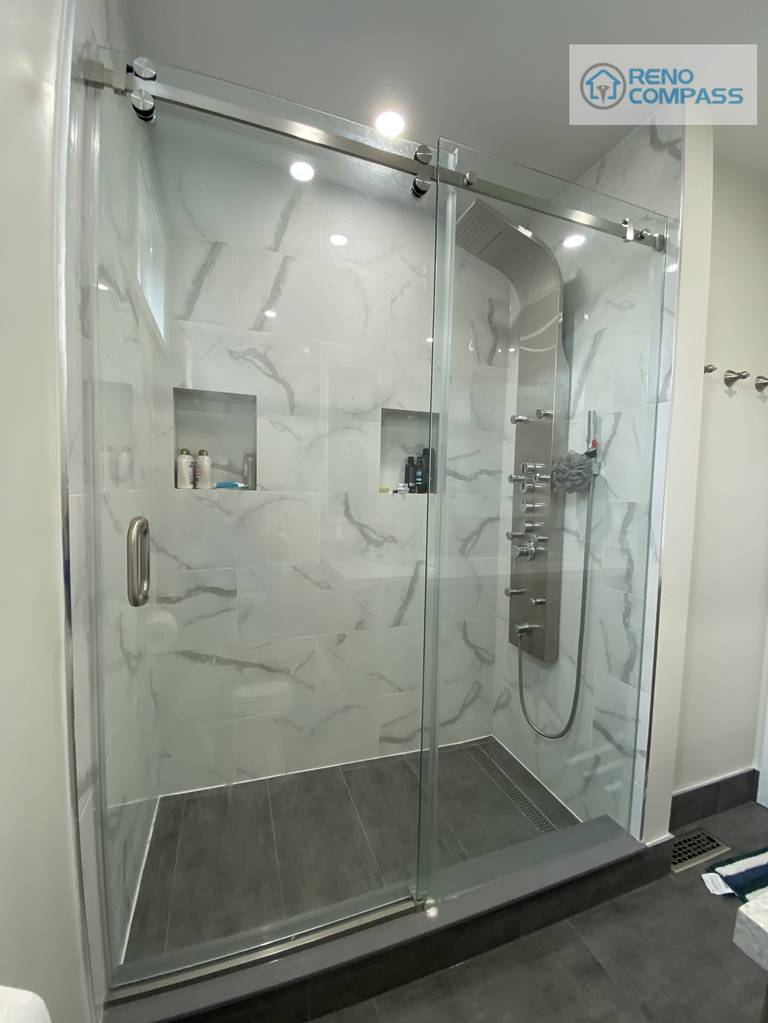 This bathroom renovation in Pickering features a frameless glass walk-in shower with marble-look wall tiles, built-in shower niches, sliding glass door system, and modern rainfall shower fixtures. The clean, hotel-style design emphasizes functionality, waterproof detailing, and contemporary aesthetics, completed by Reno Compass Inc. as part of a full interior renovation project.