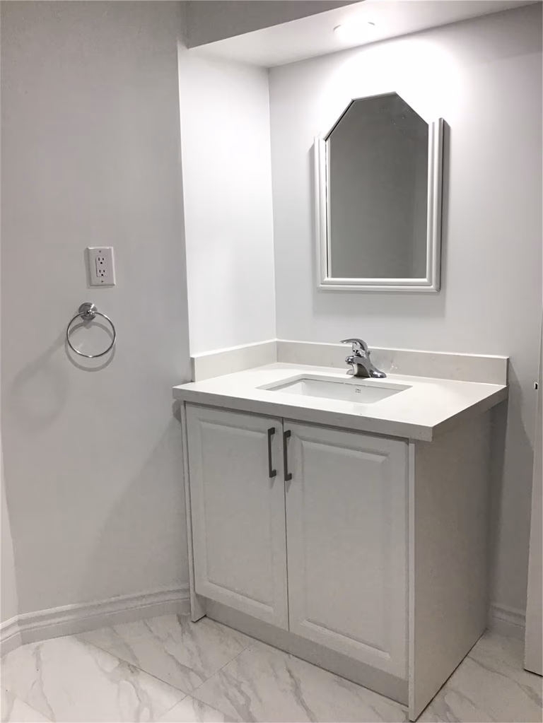 This Whitby basement renovation features a clean and modern bathroom vanity with a white integrated sink, shaker-style cabinet doors, and a framed mirror with overhead lighting. The marble-look porcelain floor tiles complement the minimalist design, creating a bright and cohesive basement bathroom space that is both functional and visually refined.