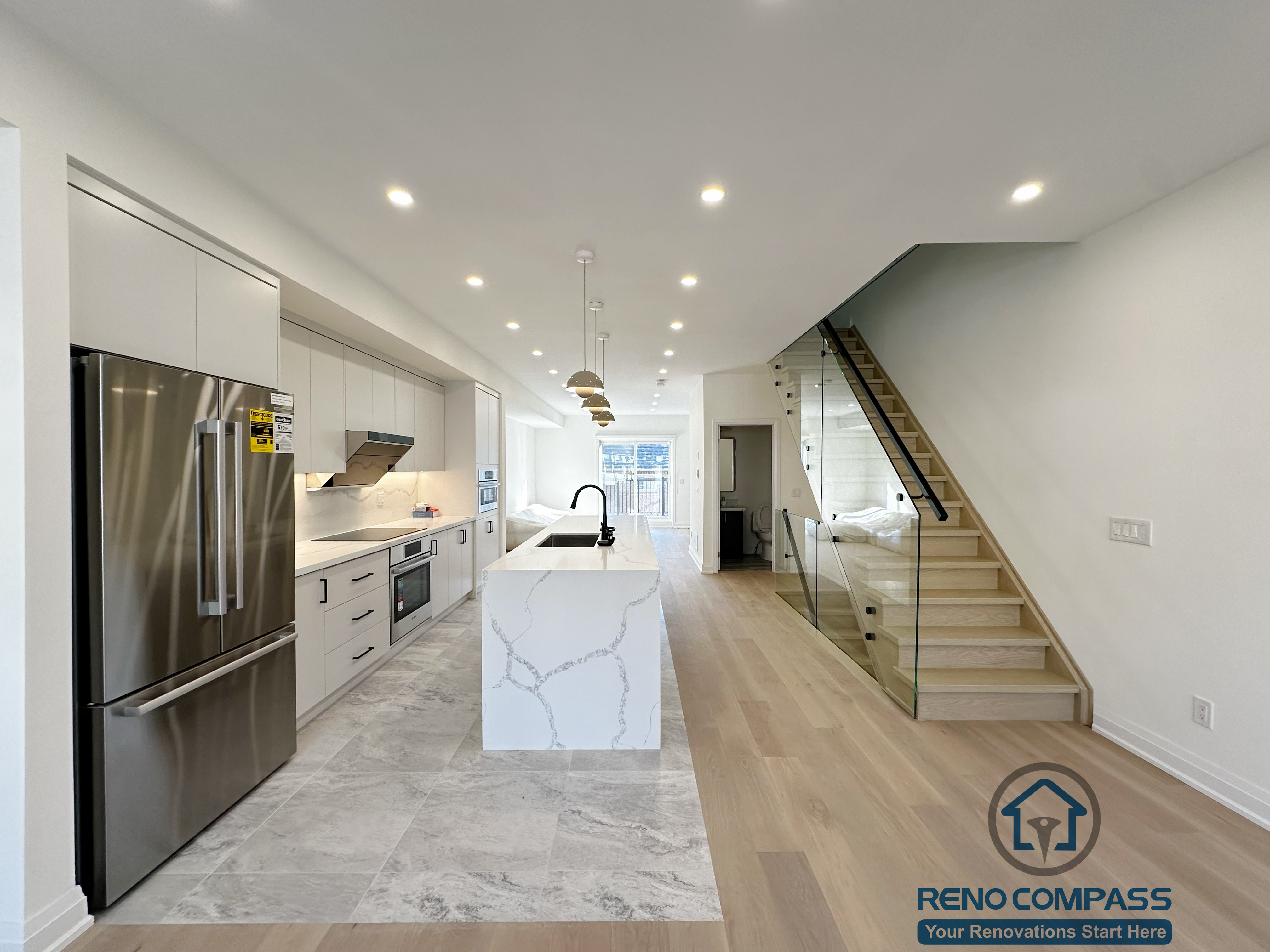 This image features a bright open-concept kitchen renovation in Richmond Hill, highlighted by a waterfall island, modern cabinetry, stainless steel appliances, and a sleek glass staircase. The clean layout, recessed lighting, and natural wood flooring create a seamless transition between kitchen, dining, and living areas.