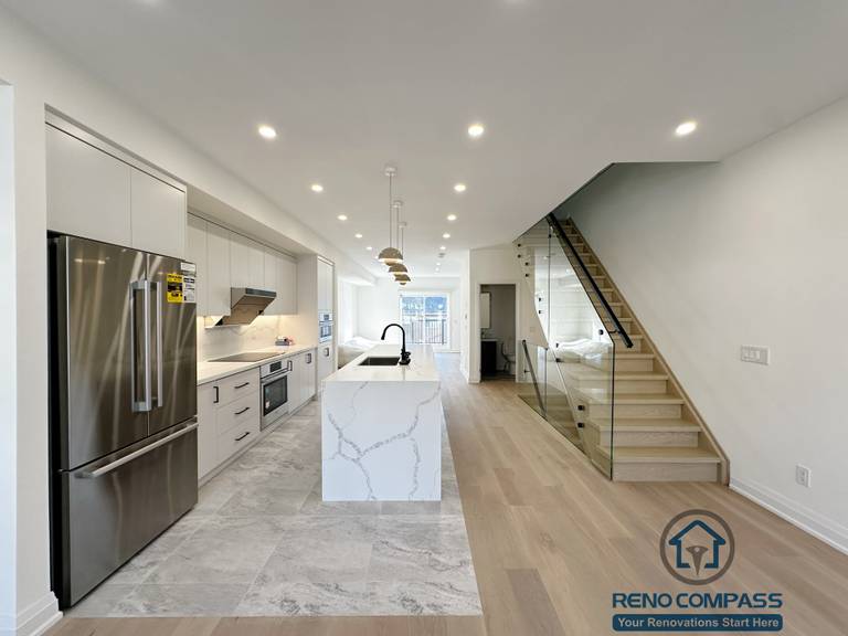 This image features a bright open-concept kitchen renovation in Richmond Hill, highlighted by a waterfall island, modern cabinetry, stainless steel appliances, and a sleek glass staircase. The clean layout, recessed lighting, and natural wood flooring create a seamless transition between kitchen, dining, and living areas.