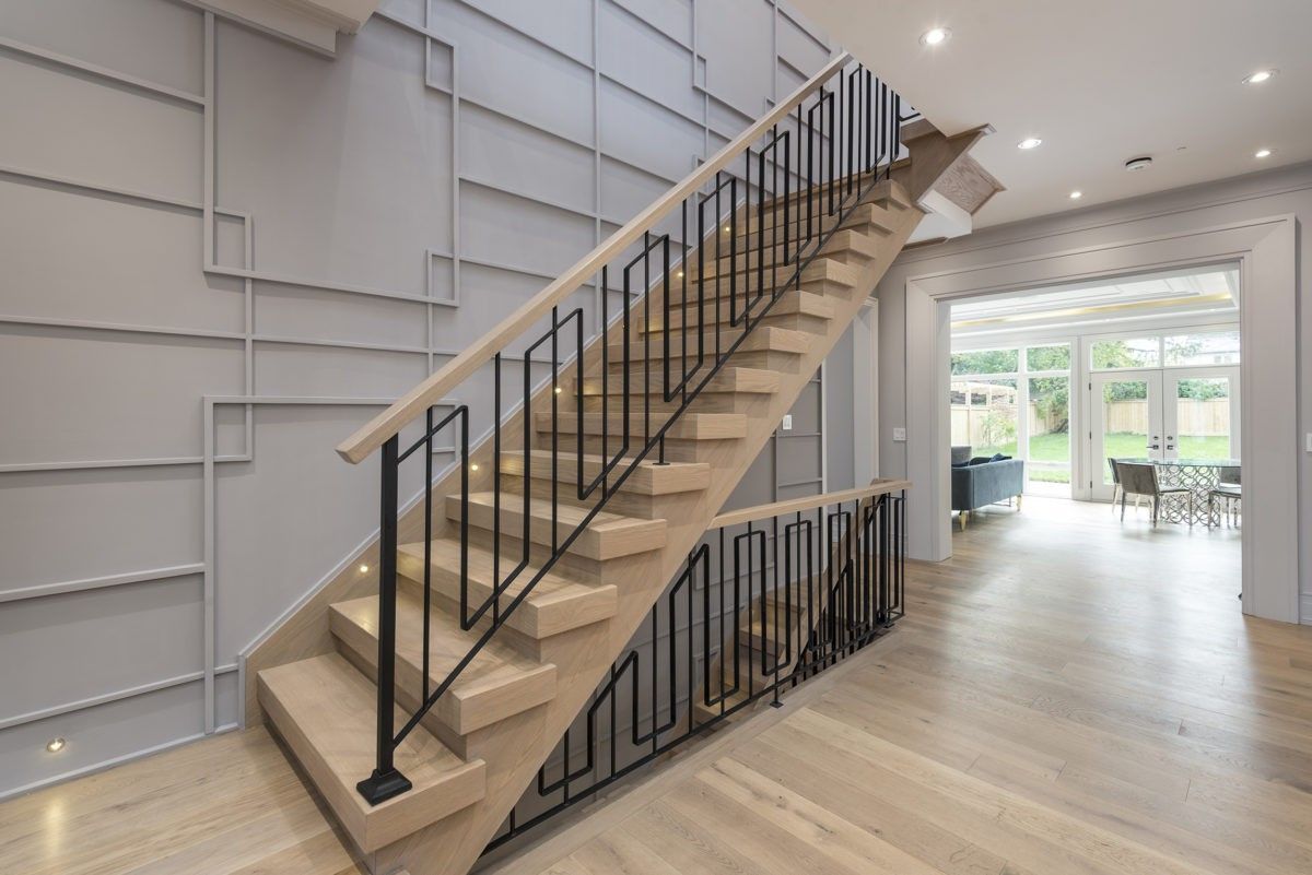 This Newmarket interior renovation showcases a custom modern staircase designed and built by a professional Toronto renovation company. The space features light oak stair treads, contemporary black metal railings, and a striking geometric feature wall that adds architectural interest and depth. Recessed lighting and wide plank hardwood flooring create a bright, open-concept atmosphere that connects seamlessly to the living and dining areas. This renovation enhances both functionality and visual impact, delivering a sophisticated modern interior for homeowners in Newmarket and across the GTA.
