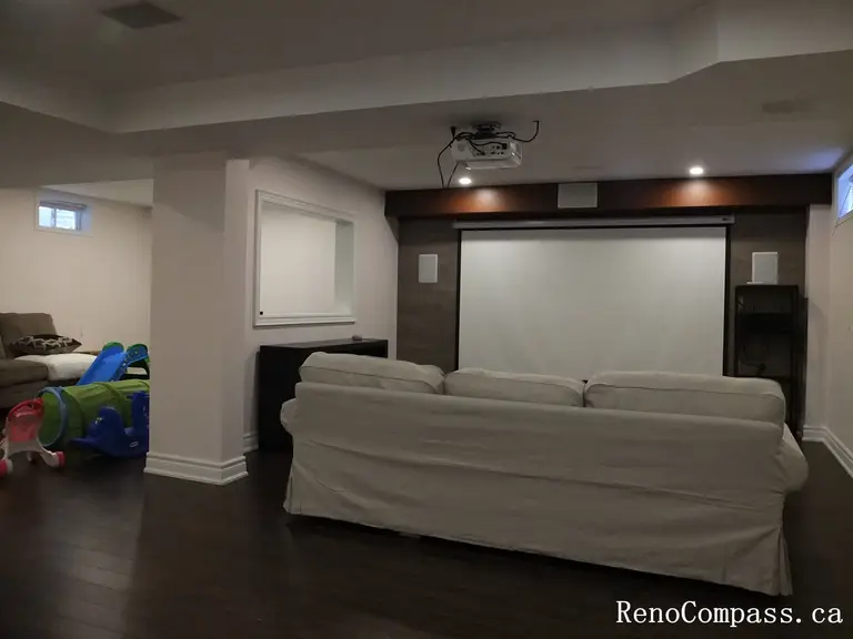 Modern basement interior renovation in Newmarket featuring a custom home theater setup, dark hardwood flooring, built-in shelving, recessed lighting, and a projector screen. Professionally completed by a Toronto Renovation Company to create a functional entertainment space for the whole family.