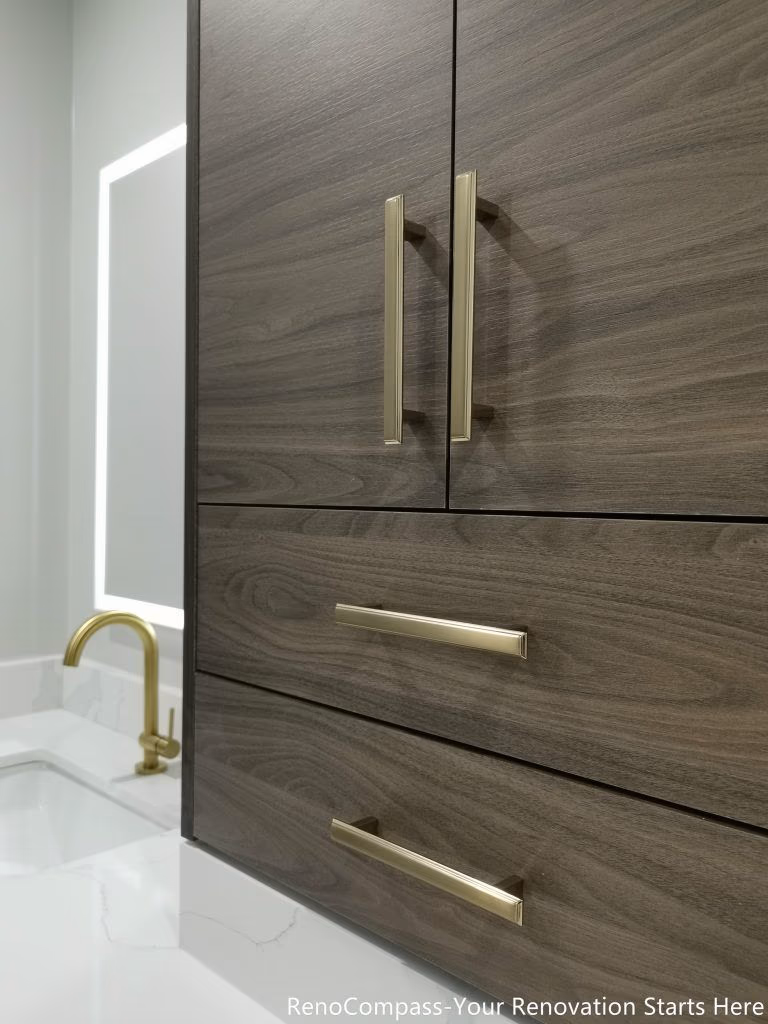 This Oakville master bathroom renovation showcases a custom-built modern vanity cabinet featuring rich wood grain finishes, quartz countertop, and elegant brushed gold hardware. The integrated storage design maximizes functionality while maintaining a clean, contemporary aesthetic. Paired with a stylish undermount sink and illuminated mirror, this upgrade enhances both daily convenience and visual appeal. Completed by a trusted Toronto renovation company, this bathroom vanity installation delivers a perfect balance of luxury, durability, and practical storage solutions.