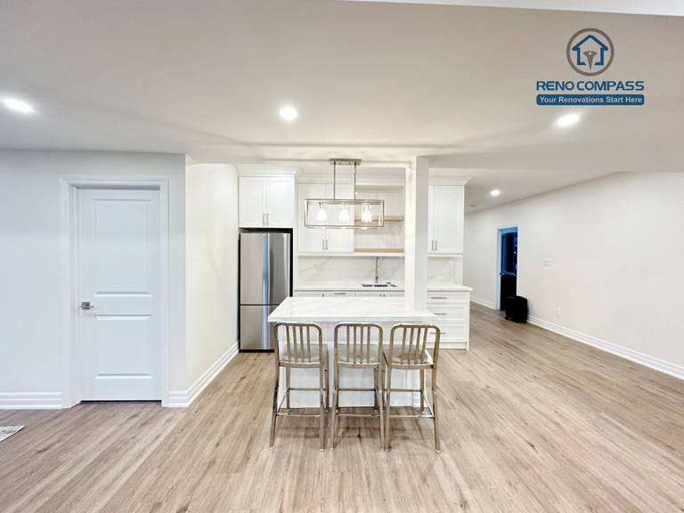 This Bradford basement renovation transforms the lower level into a bright, open-concept living and entertainment space. The layout integrates a custom wet bar with full-height cabinetry, quartz countertop, and built-in sink, creating a functional transition between the entertainment zone and seating area. Recessed LED lighting throughout the ceiling enhances brightness and eliminates the typical basement dark feel, while durable wood-look flooring provides warmth and continuity across the space. Structural columns were carefully incorporated into the design to maintain openness without compromising support. This basement renovation reflects Reno Compass’ approach to practical space planning, clean finishes, and long-term usability for modern family living.
