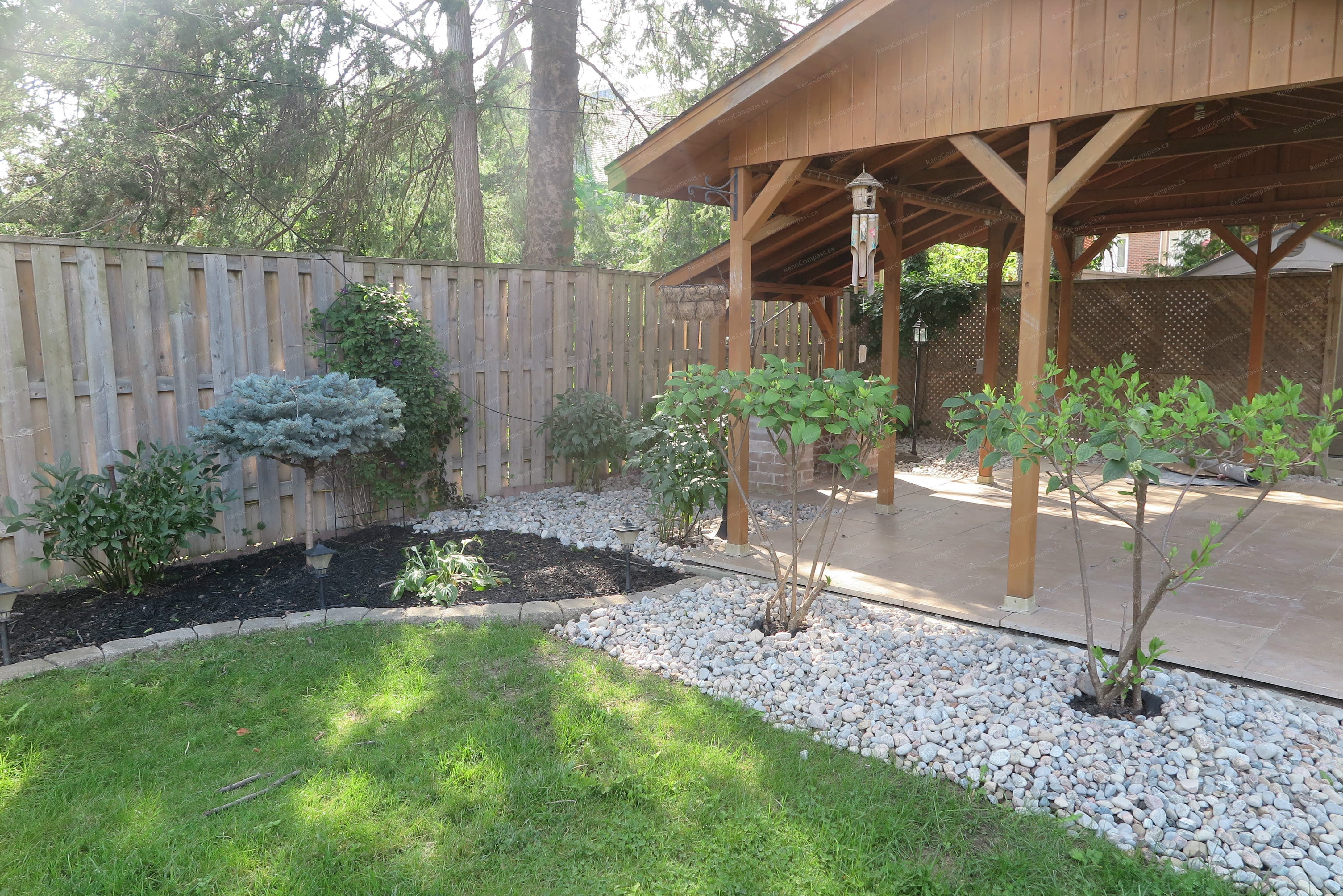 This image highlights a North York outdoor landscaping renovation featuring a wooden pavilion, decorative stone borders, and layered garden beds. The design creates a seamless connection between lawn, planting areas, and covered seating space while improving drainage and long-term maintenance. Completed by a Toronto Renovation Company specializing in custom backyard landscaping and outdoor structure integration.