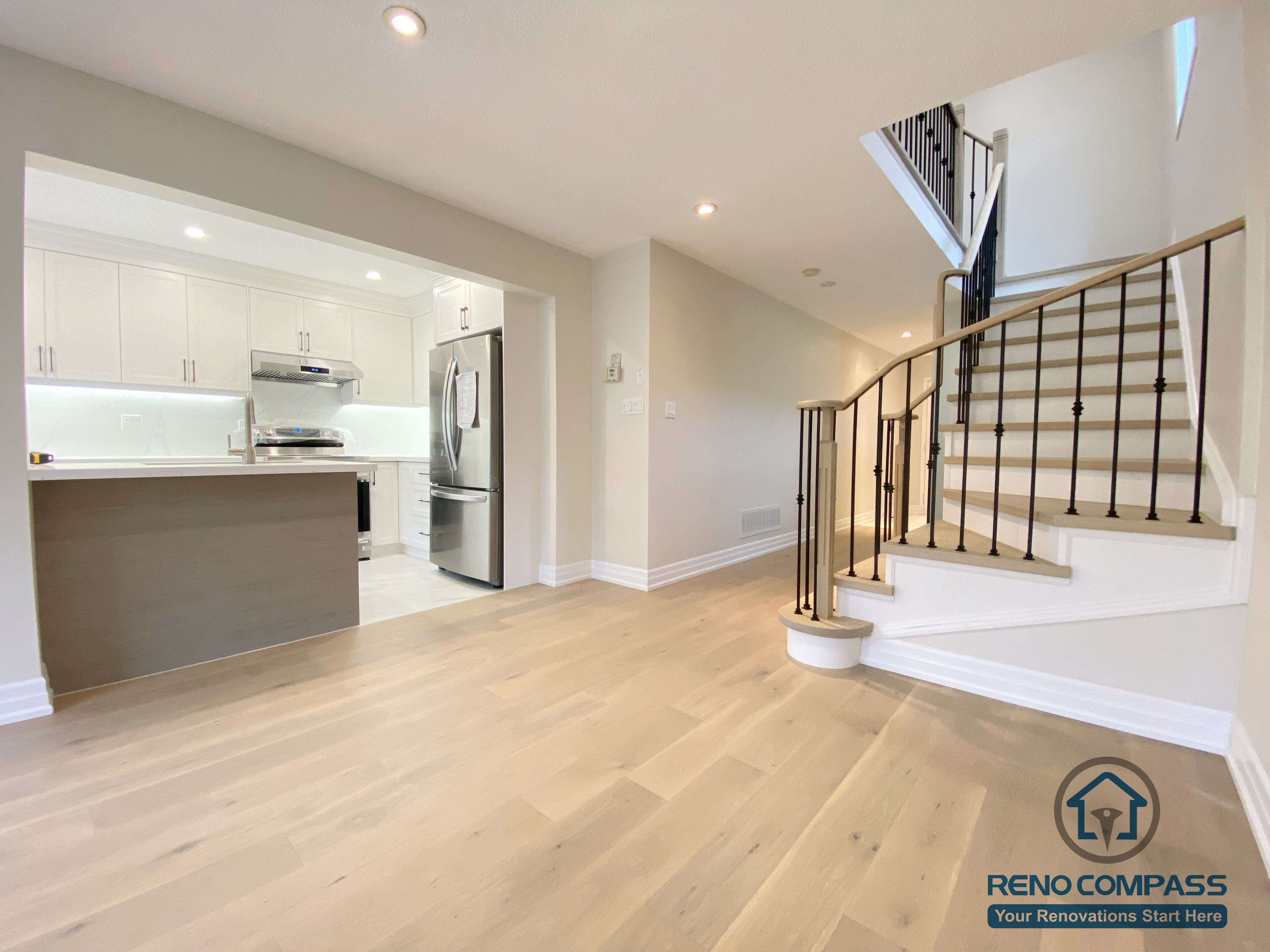 This Markham interior renovation showcases an open-concept layout that seamlessly connects the kitchen, living area, and staircase. The space features wide-plank hardwood flooring, a modern kitchen with white cabinetry and quartz countertops, and a refined staircase with hardwood treads and black metal railings. Recessed lighting enhances brightness and continuity throughout the main floor. Completed by a professional Toronto renovation company, this design improves circulation, visual balance, and everyday functionality for a Markham home.