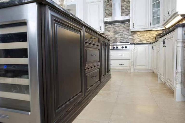 This European-style kitchen renovation showcases a custom dark wood island with detailed panel molding and integrated storage drawers, creating a bold contrast against the surrounding white cabinetry. The island features a built-in wine cooler, adding both convenience and upscale functionality to the space.

The natural stone countertop enhances durability and elegance, while the textured stone backsplash and stainless steel range hood provide a refined focal point in the background. Neutral tile flooring completes the cohesive, high-end design.

This layout highlights premium craftsmanship and thoughtful appliance integration, ideal for homeowners seeking a luxury kitchen renovation with custom millwork and functional island features.