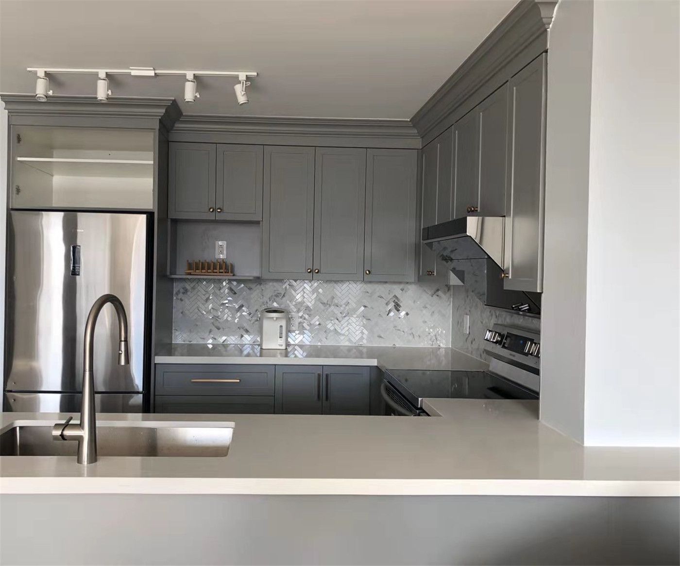 Contemporary condo kitchen renovation in Richmond Hill featuring elegant grey shaker cabinets, a bright quartz countertop, and stainless steel appliances. The space is enhanced with a stylish herringbone tile backsplash, modern track lighting, and a functional kitchen layout that maximizes storage and cooking efficiency while creating a clean and sophisticated look.
