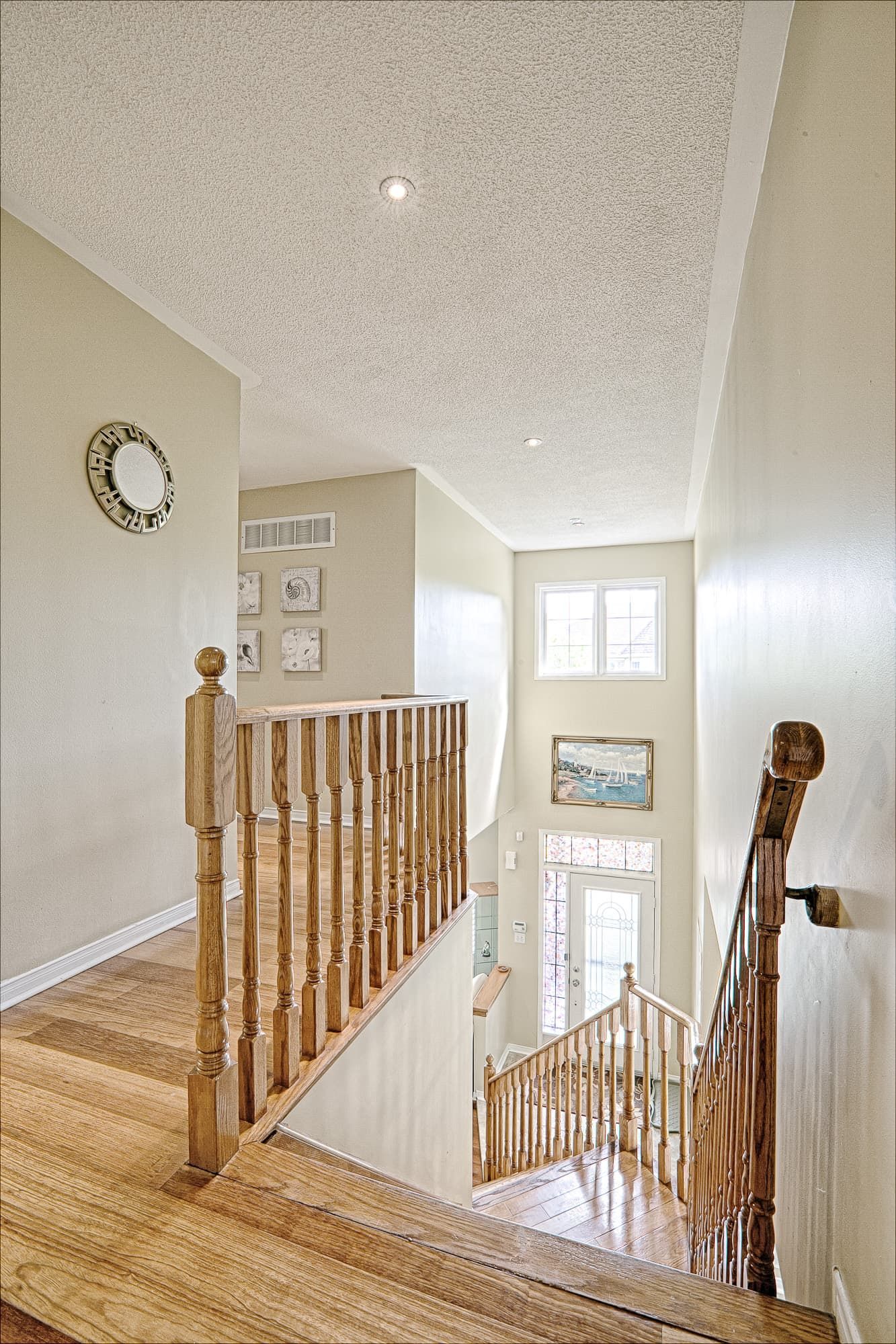 This bright staircase and upper hallway design showcases natural hardwood flooring, classic wooden railings, and soft neutral wall tones that enhance openness and flow between levels. The double-height space allows natural light to fill the stairwell, creating a clean and welcoming transition area within the home. An excellent example of warm minimalist interior renovation and functional home design inspiration for homeowners in Toronto and the GTA.