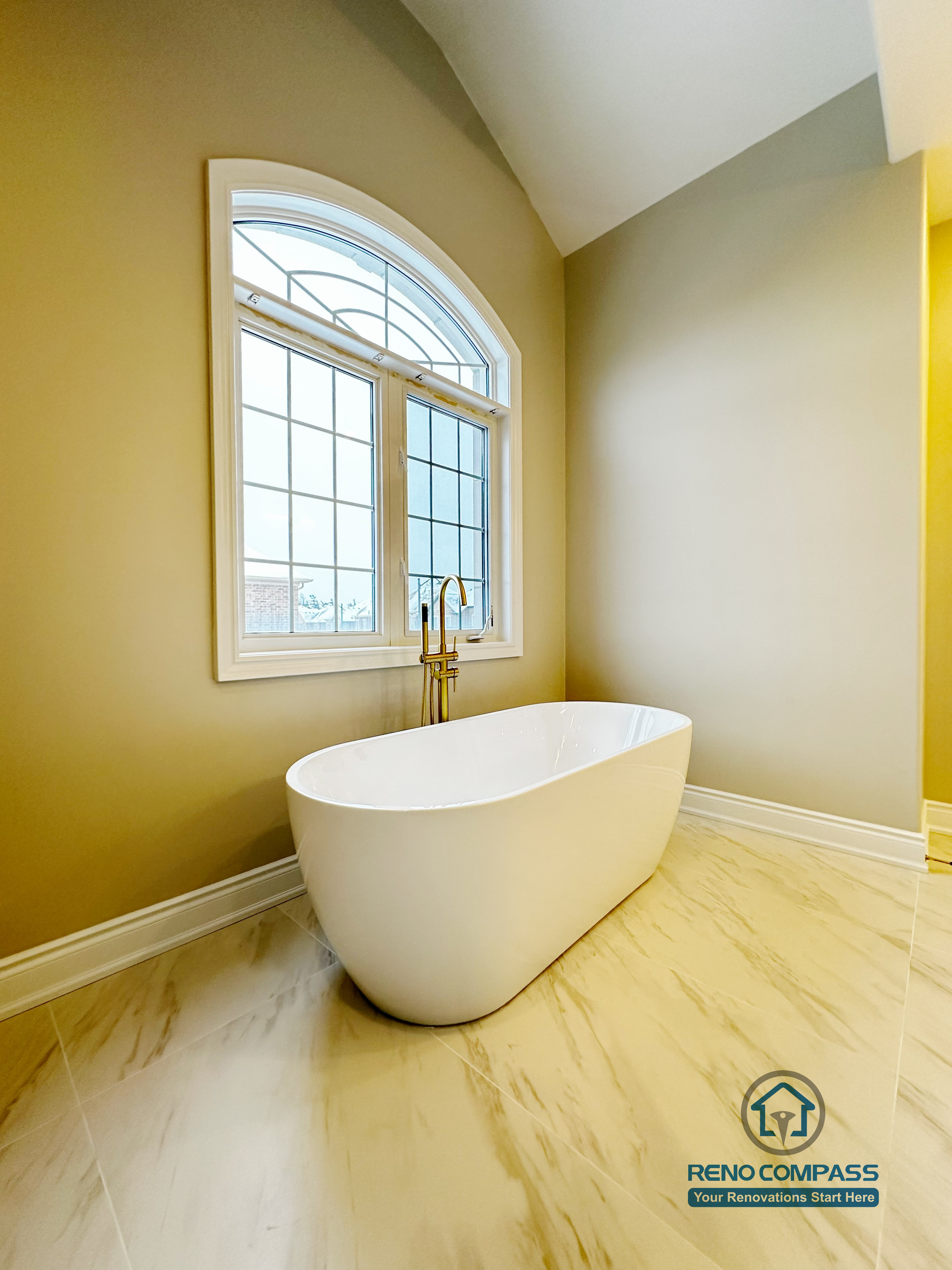 Luxury freestanding soaking tub featured in a Vaughan primary bathroom renovation, positioned beneath an elegant arched window for natural light. Finished with marble-look porcelain flooring and a modern floor-mounted tub filler, this spa-inspired space was designed by a professional Toronto renovation company to enhance comfort, openness, and relaxation.
