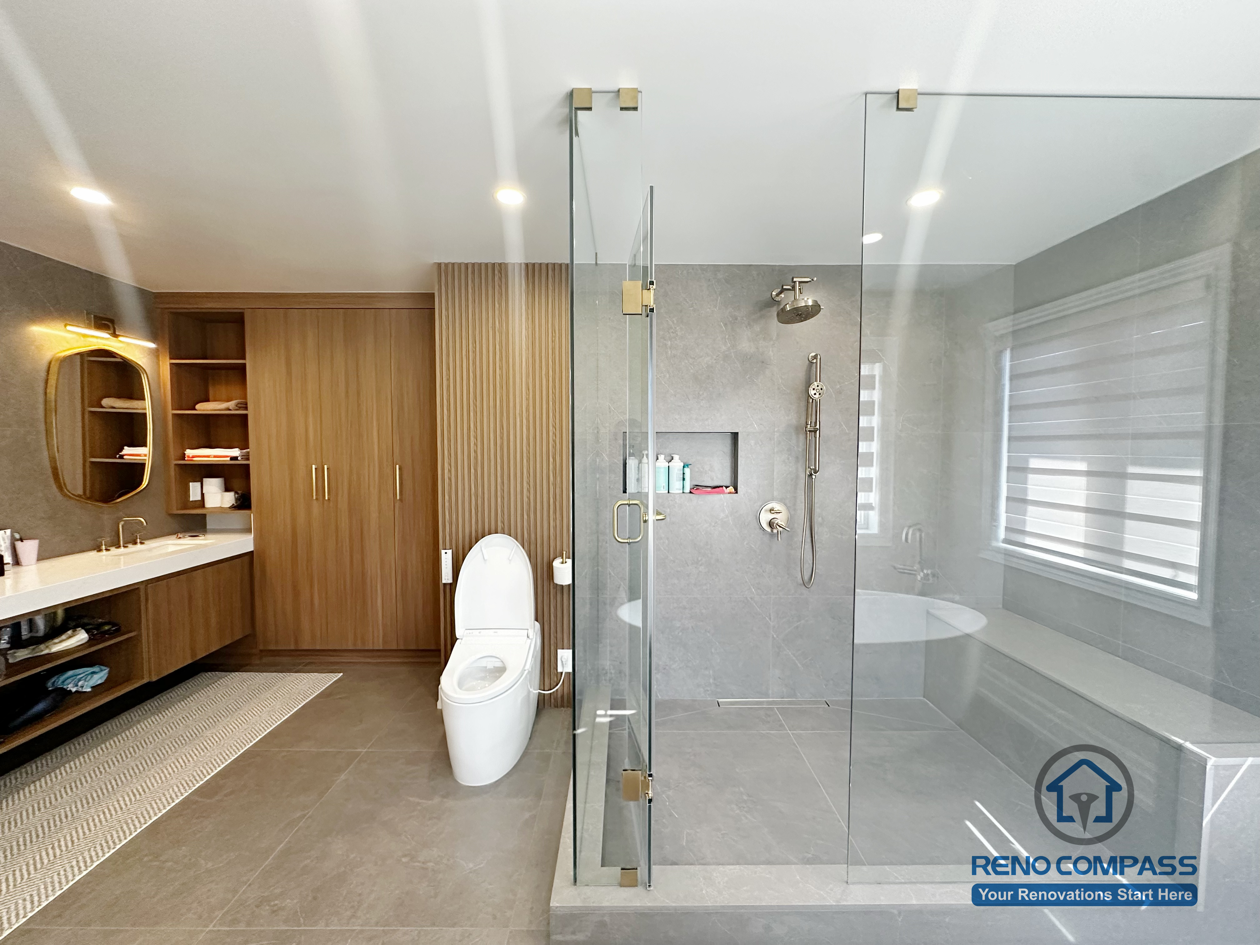 This image showcases a spacious primary bathroom renovation completed in North York, featuring a walk-in glass shower with built-in niche, a freestanding soaking tub, and custom wood storage cabinetry. Large-format floor tiles provide durability and visual continuity, while the frameless shower enclosure enhances openness and natural light. The layout balances functionality and comfort, creating a modern bathroom designed for everyday use and long-term living in North York homes.
