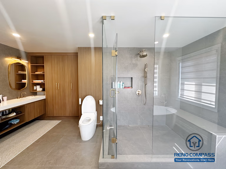 This image showcases a spacious primary bathroom renovation completed in North York, featuring a walk-in glass shower with built-in niche, a freestanding soaking tub, and custom wood storage cabinetry. Large-format floor tiles provide durability and visual continuity, while the frameless shower enclosure enhances openness and natural light. The layout balances functionality and comfort, creating a modern bathroom designed for everyday use and long-term living in North York homes.
