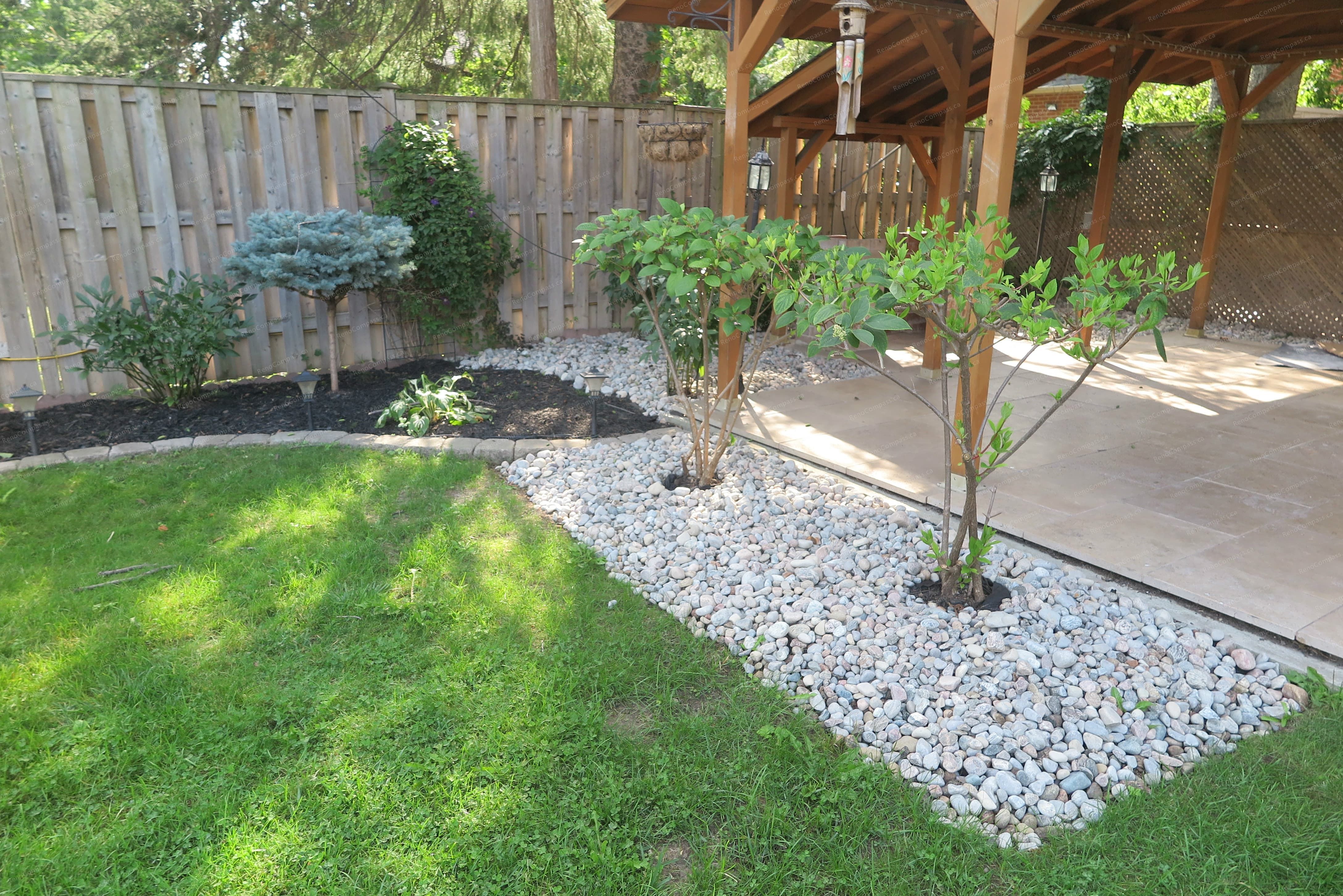 North York outdoor landscaping project showcasing decorative white gravel garden beds, newly planted shrubs, natural lawn area, and a custom wooden pavilion. Designed and built by Reno Compass to create a clean, low-maintenance backyard with a relaxing outdoor living and garden space.