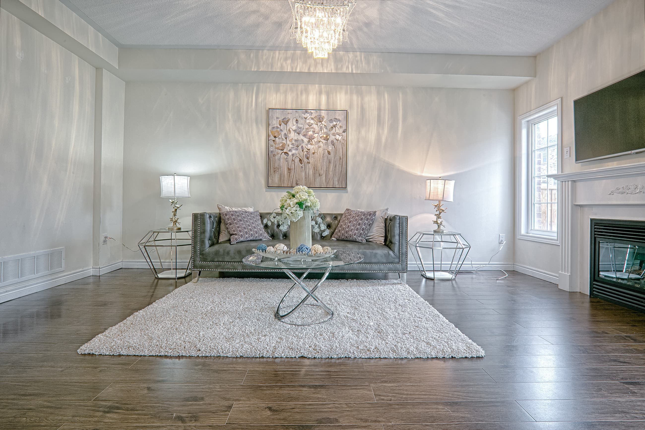 This modern chic living room renovation showcases a bright open layout with elegant crystal chandelier lighting, dark hardwood flooring, and a refined neutral color palette. The space features contemporary furniture styling, a textured area rug, and a sleek fireplace wall that creates a balanced blend of comfort and luxury. Designed with clean lines and sophisticated finishes, this interior highlights the craftsmanship of a professional Toronto renovation company delivering high-quality full home interior renovations tailored for modern family living.