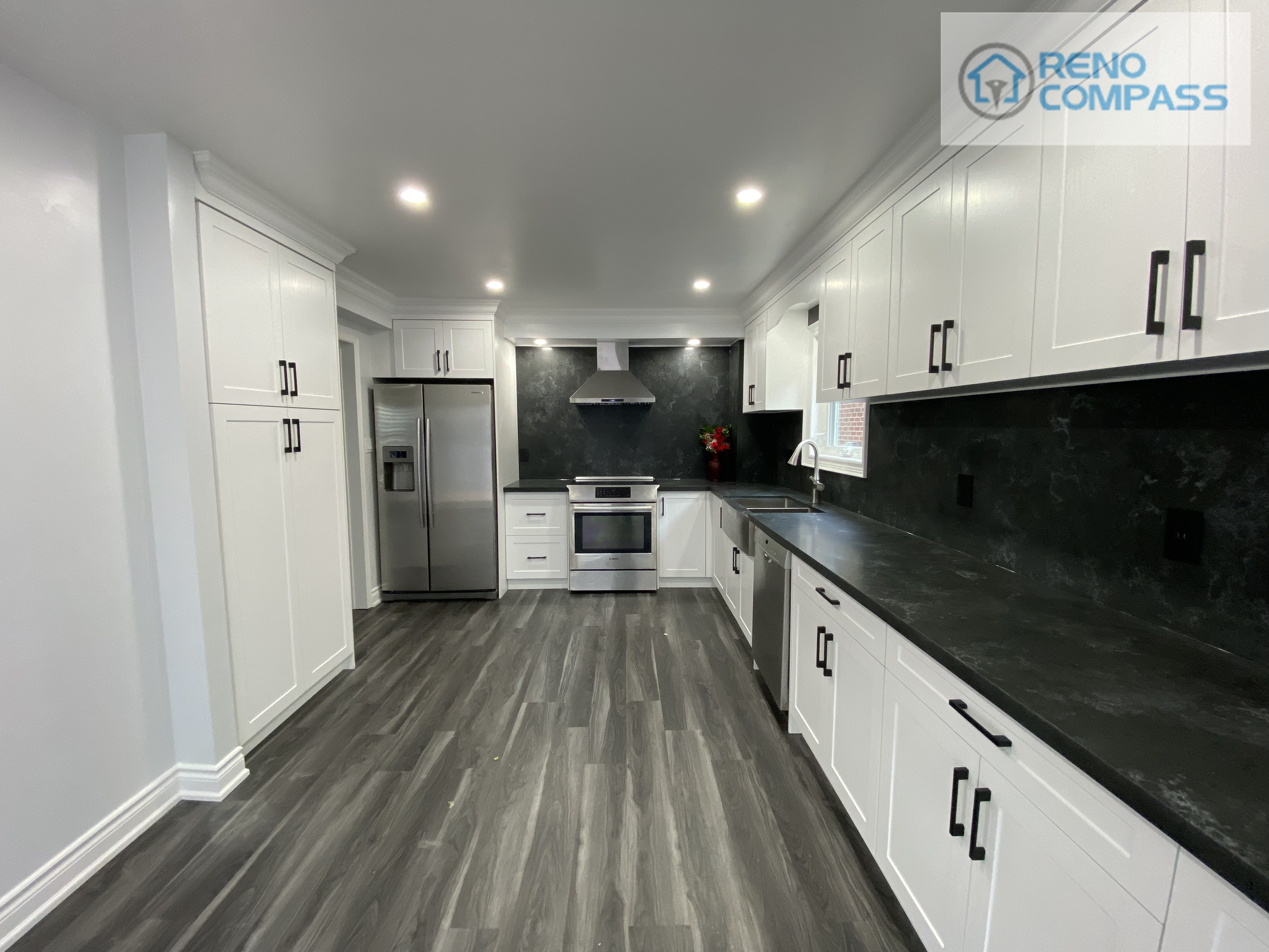 Wide-angle view of a modern kitchen interior renovation in Pickering, featuring full-height white shaker cabinetry, black stone countertops and backsplash, stainless steel appliances, recessed ceiling lighting, and durable vinyl plank flooring. This clean and functional kitchen layout was designed and built by Reno Compass Inc., delivering a bright, streamlined space optimized for everyday use.