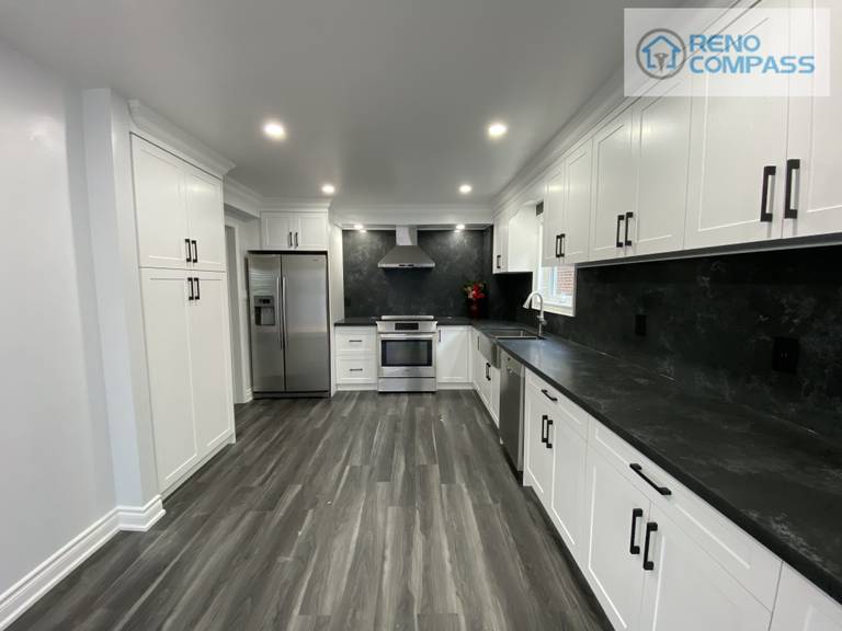 Wide-angle view of a modern kitchen interior renovation in Pickering, featuring full-height white shaker cabinetry, black stone countertops and backsplash, stainless steel appliances, recessed ceiling lighting, and durable vinyl plank flooring. This clean and functional kitchen layout was designed and built by Reno Compass Inc., delivering a bright, streamlined space optimized for everyday use.