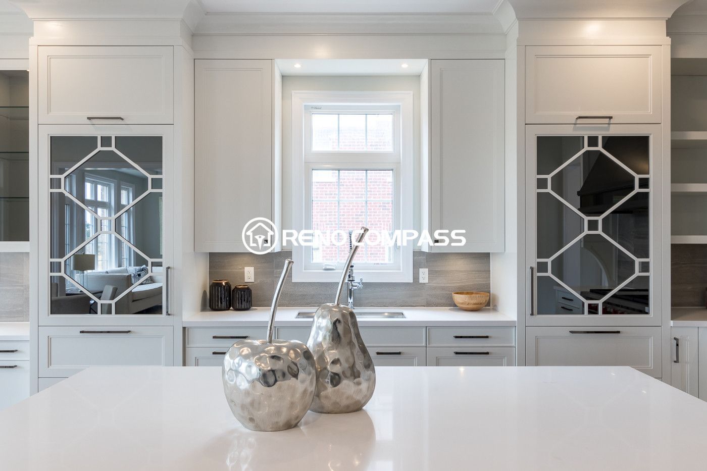 This elegant Newmarket kitchen renovation features custom white cabinetry, a centered window above the sink, and a spacious quartz countertop island. Decorative glass cabinet panels and soft lighting add a refined and modern aesthetic to the space. Completed by a trusted Toronto renovation company, Reno Compass Inc., the design combines functionality with timeless kitchen craftsmanship.