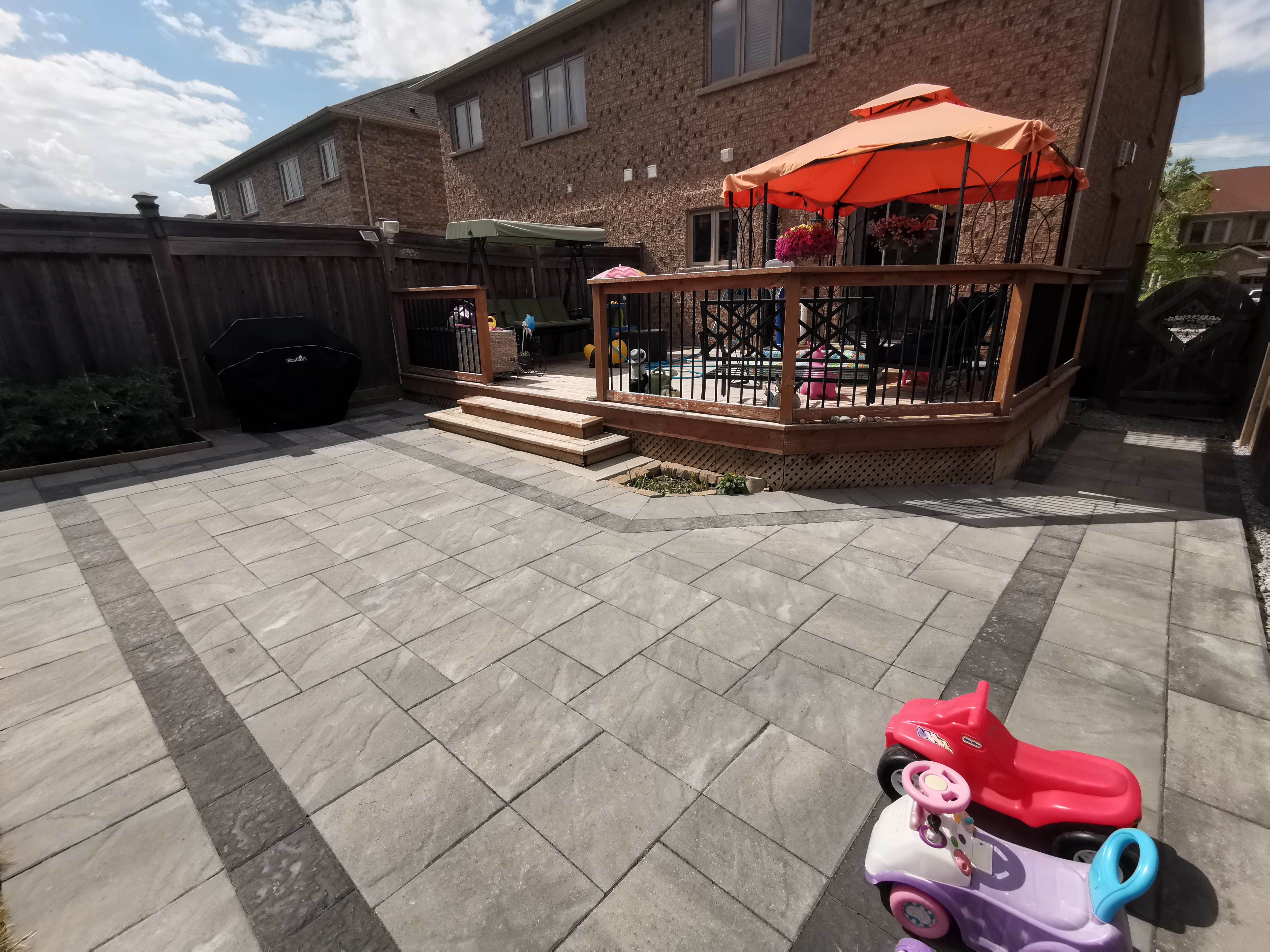 This Aurora backyard interlocking paver patio is designed to create a functional and inviting outdoor living space. Large-format concrete pavers are installed in a clean linear pattern with a contrasting border, enhancing visual definition and long-term durability. The patio seamlessly connects to a raised wooden deck with integrated steps, allowing smooth movement between indoor and outdoor areas. This professionally built interlock patio offers excellent drainage, stability, and low maintenance, making it ideal for family use and everyday enjoyment in residential backyards across Aurora.