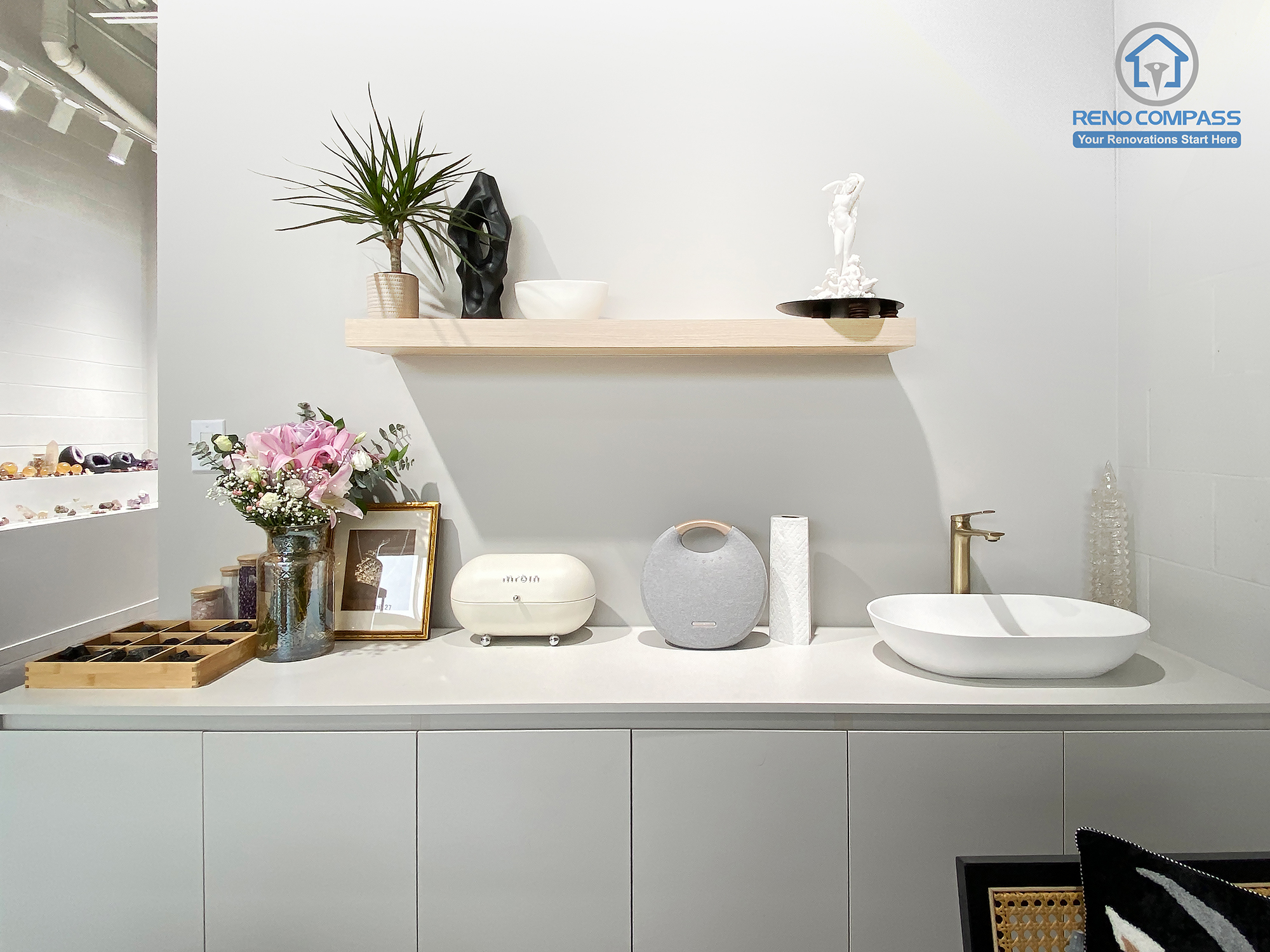 This image features a custom cabinetry and utility counter designed as part of a crystal shop renovation in Markham. The space includes built-in storage cabinets, a minimalist countertop with integrated sink, floating display shelf, and carefully styled decorative elements. Soft neutral finishes and functional layout support daily retail operations while maintaining a calm, curated aesthetic. As a Toronto renovation company, Reno Compass delivers retail renovation solutions that combine practical workspace design with refined visual presentation.