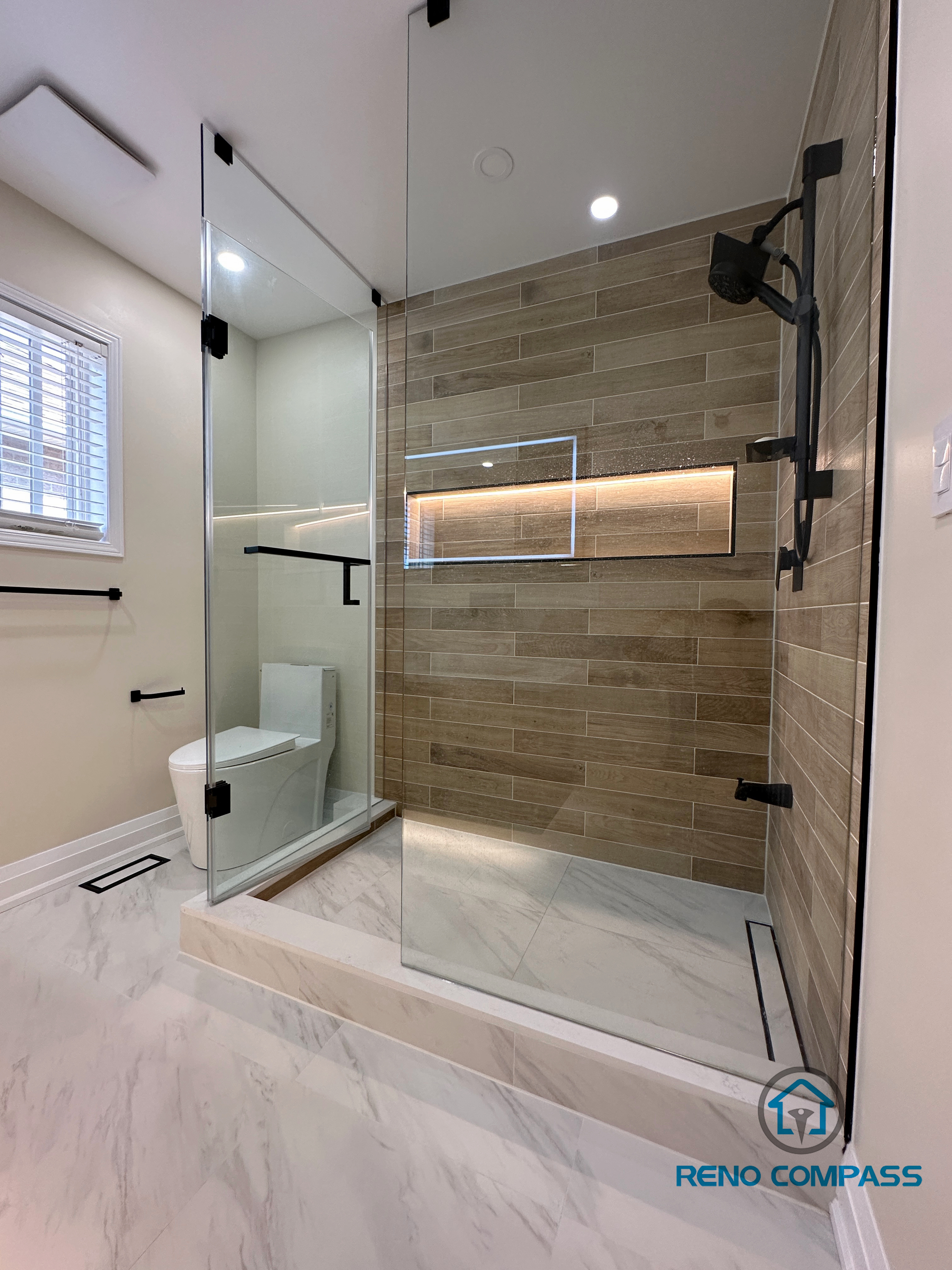 Contemporary bathroom renovation in Vaughan featuring a spacious walk-in shower with a frameless glass enclosure, custom LED-lit shower niche, and wood-look porcelain wall tiles. The shower design includes matte black fixtures, a low-profile shower base, and marble-look porcelain flooring for a seamless and modern finish. This project demonstrates high-quality workmanship and thoughtful layout planning by a trusted Toronto renovation company specializing in custom bathroom renovations.
