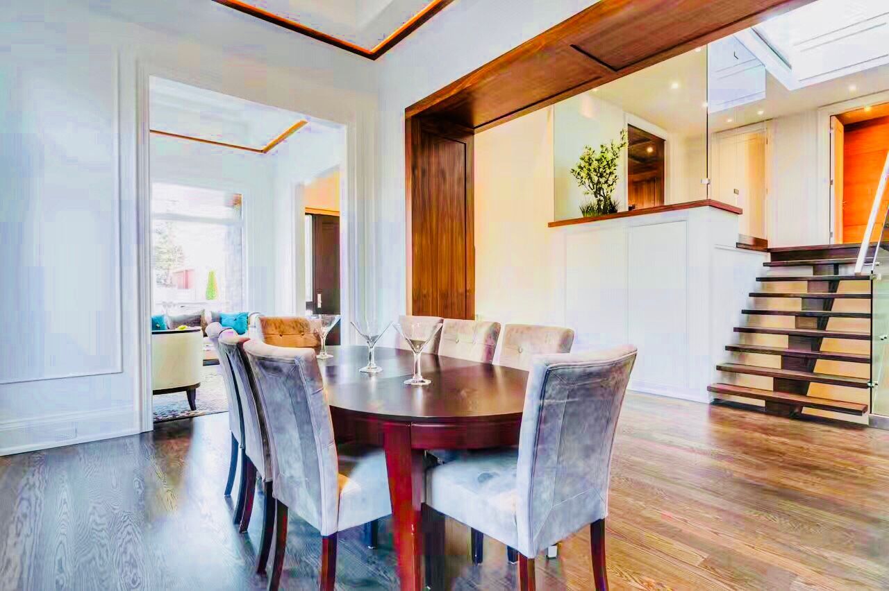 Interior view showcasing the seamless transition between the formal dining area and the main living space. Features include dark oak hardwood flooring, designer velvet dining chairs, and a contemporary glass-and-steel staircase that maximizes the sense of space in this luxury Toronto custom build.