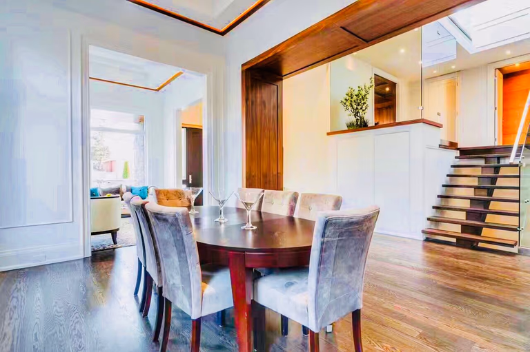 Interior view showcasing the seamless transition between the formal dining area and the main living space. Features include dark oak hardwood flooring, designer velvet dining chairs, and a contemporary glass-and-steel staircase that maximizes the sense of space in this luxury Toronto custom build.