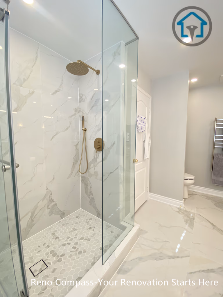 This Mississauga bathroom renovation features a spacious walk-in shower enclosed with frameless clear glass panels to enhance openness and natural light. Large-format marble-look wall tiles are paired with a mosaic tile shower floor for slip resistance and visual contrast. Gold-tone shower fixtures add warmth and elegance, while a built-in wall niche provides functional storage without disrupting the clean layout. The shower design prioritizes comfort, drainage efficiency, and long-term durability through precise detailing and quality materials.